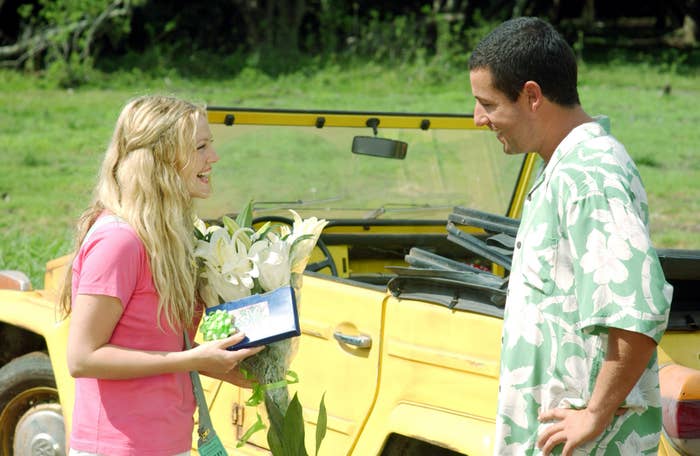 A woman holds flowers and a card, smiling at a man in a floral shirt, standing beside a yellow jeep in a grassy area