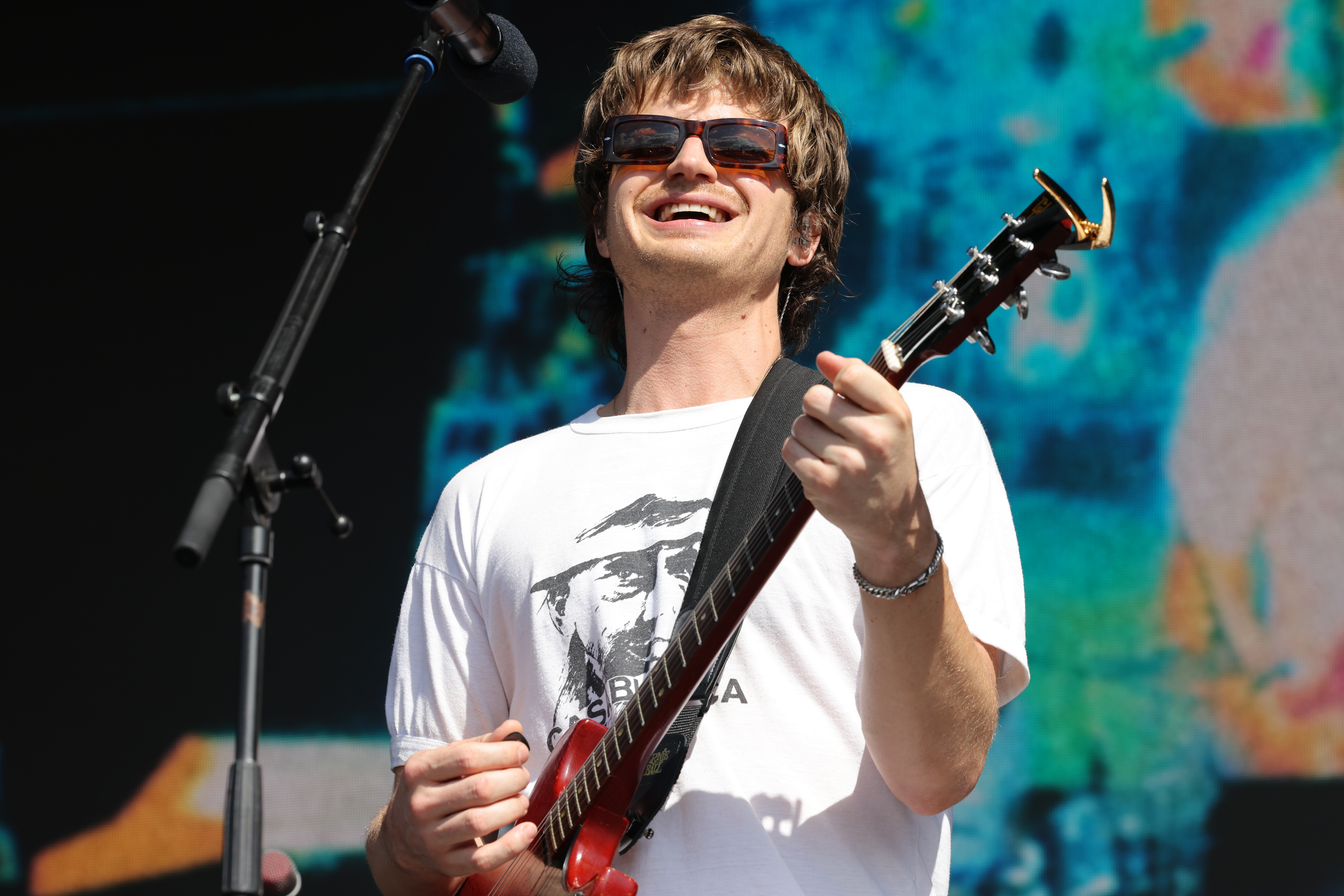 Musician performs onstage, wearing sunglasses and a graphic t-shirt, smiling while playing an electric guitar. Microphone and colorful backdrop visible
