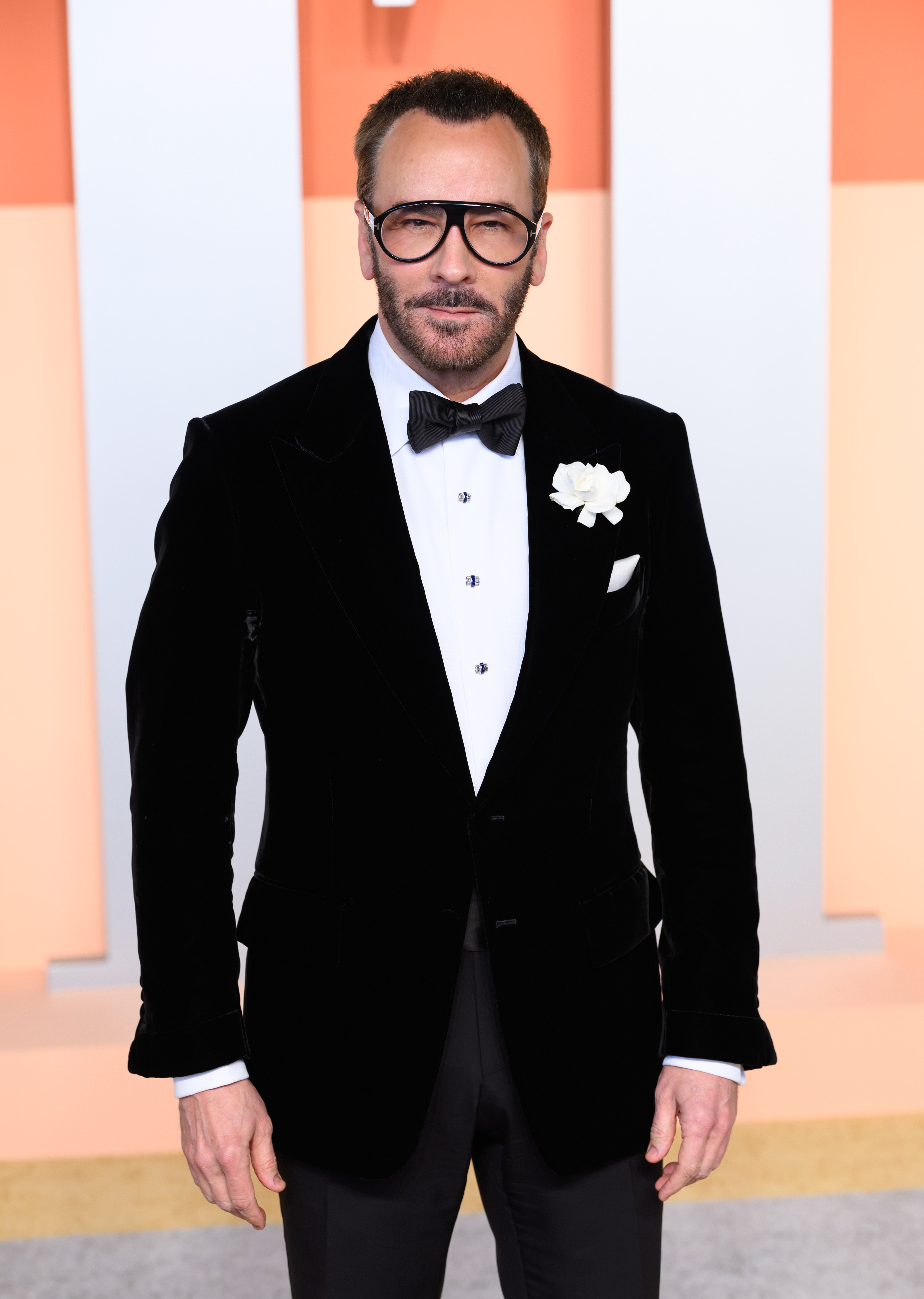 Man in a velvet tuxedo with bow tie, large glasses, and a white flower lapel pin on a red carpet