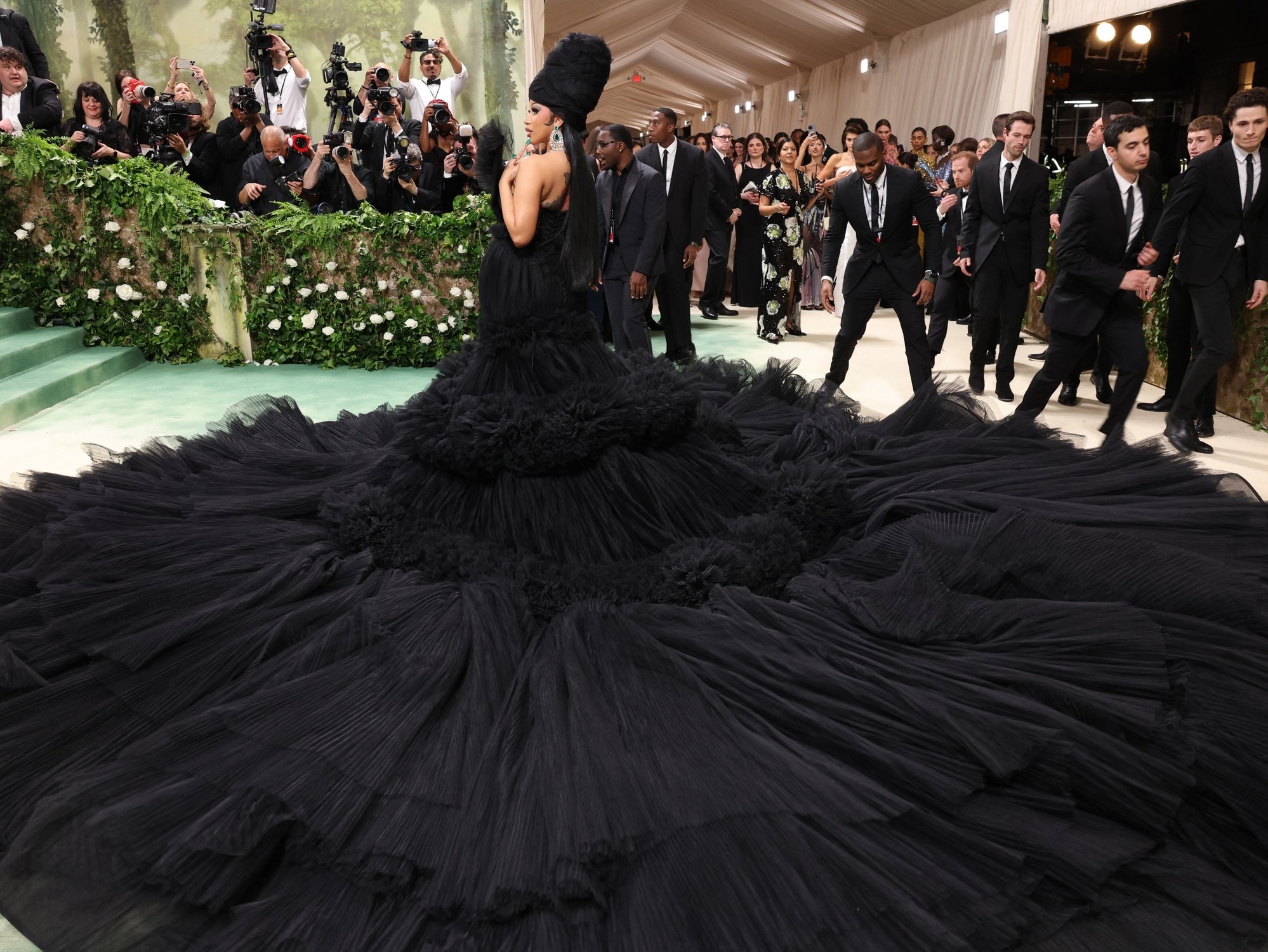 Persona con un vestido de gala voluminoso en la alfombra roja, rodeada de fotógrafos y asistentes. Estilo elegante y dramático