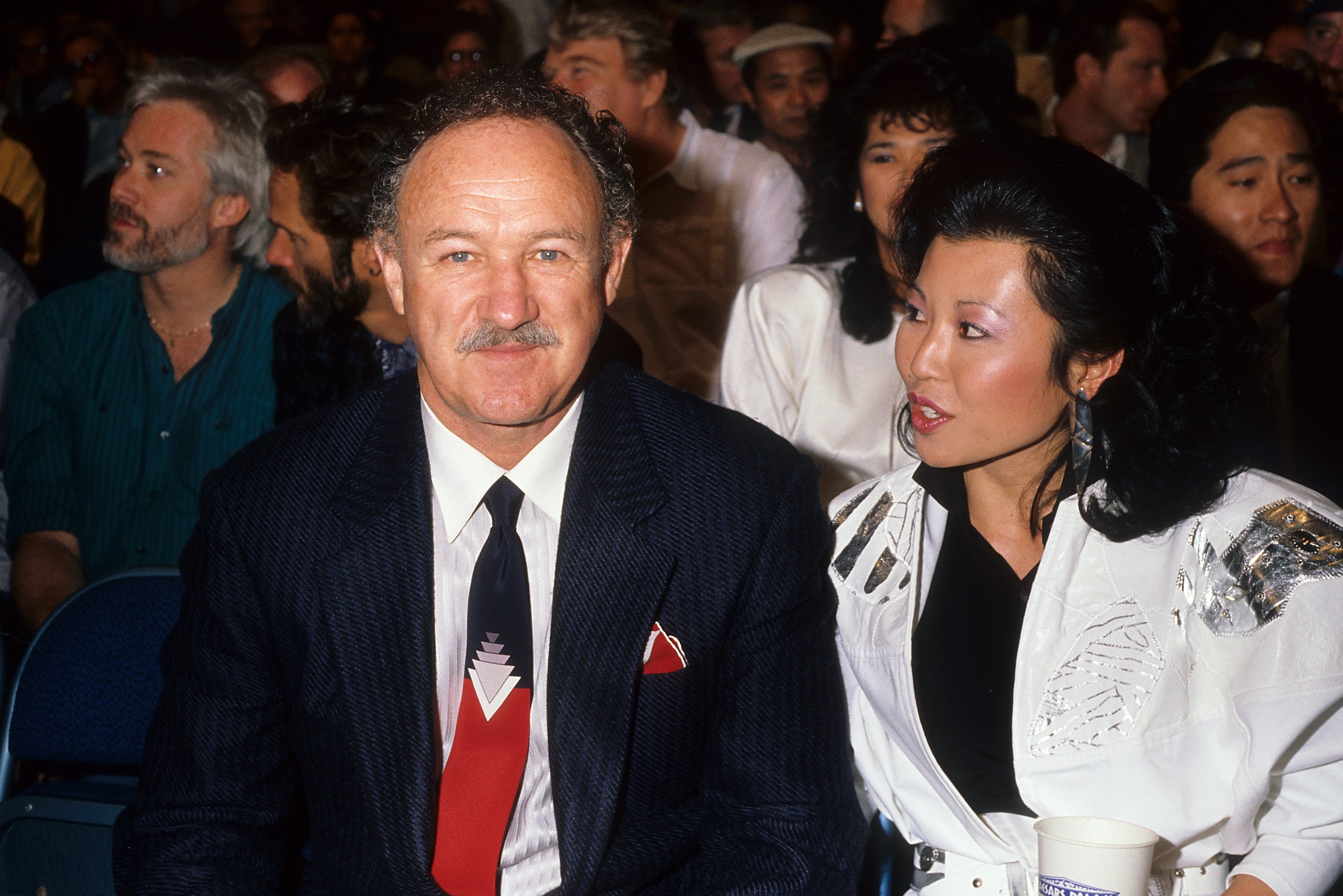 Man in a suit with a tie and woman in a stylish white outfit at an indoor event, surrounded by a crowd