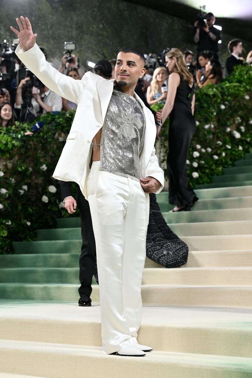 Man in white suit and shimmering top waves on a formal staircase, attended by photographers and onlookers in evening attire
