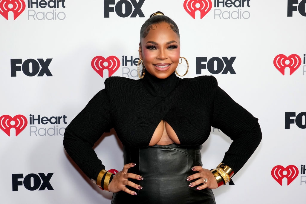 Person in a stylish black outfit with bold gold bracelets poses at an iHeartRadio and FOX event backdrop