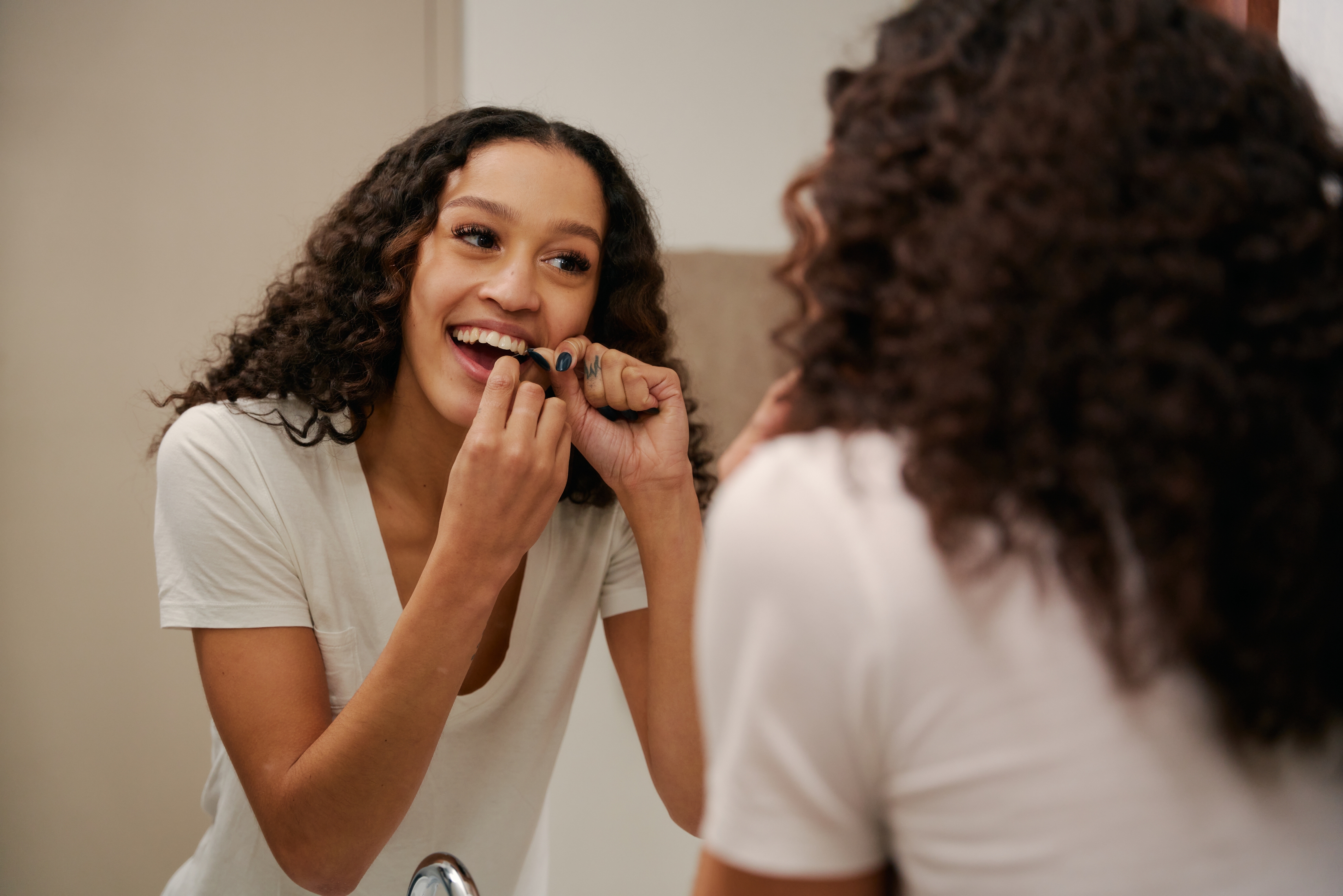 En kvinne smiler mens hun flosser tennene foran et baderomsspeil