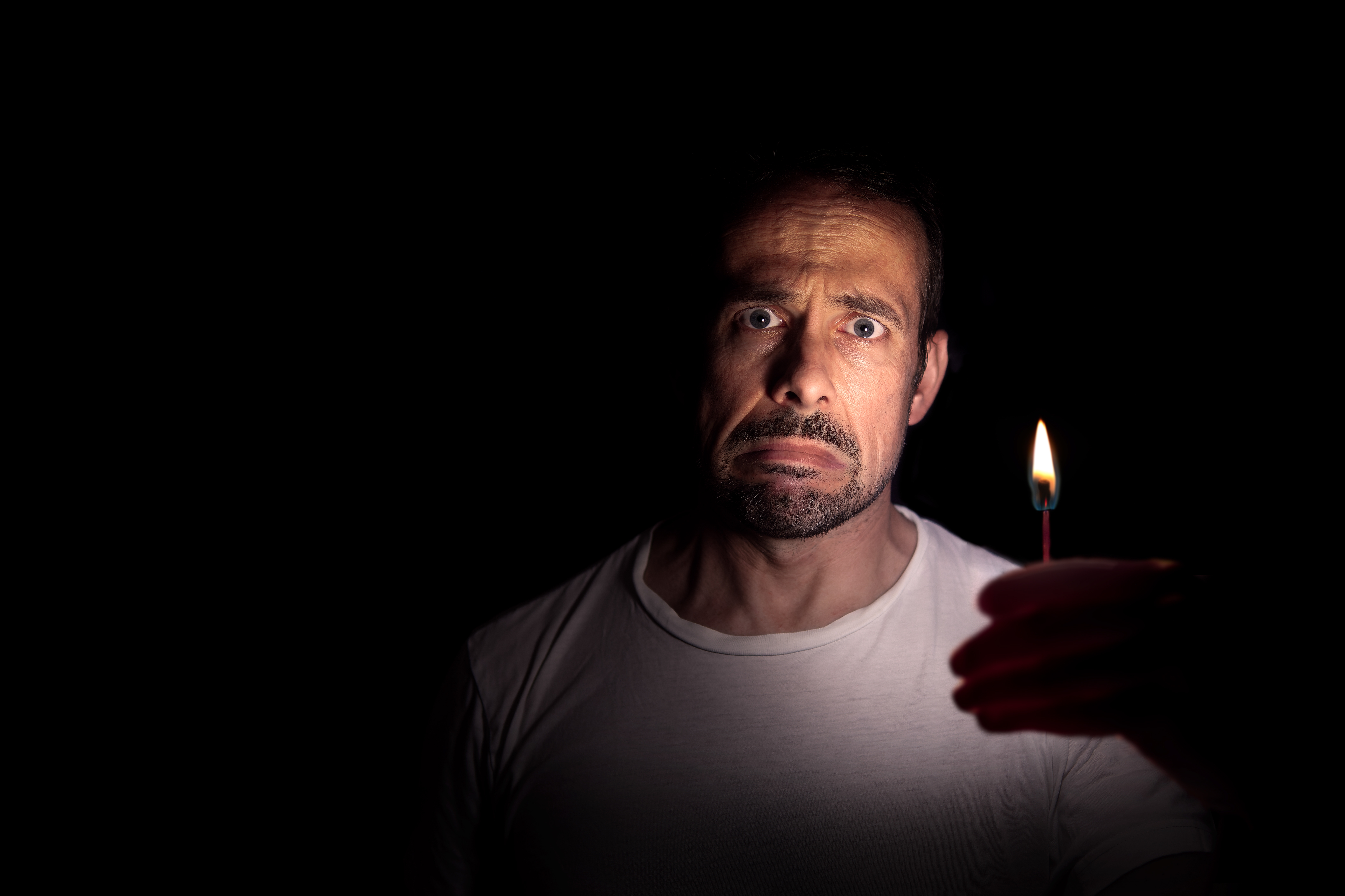 Person in a white t-shirt looks concerned, holding a lit match in the dark, casting dramatic shadows on their face