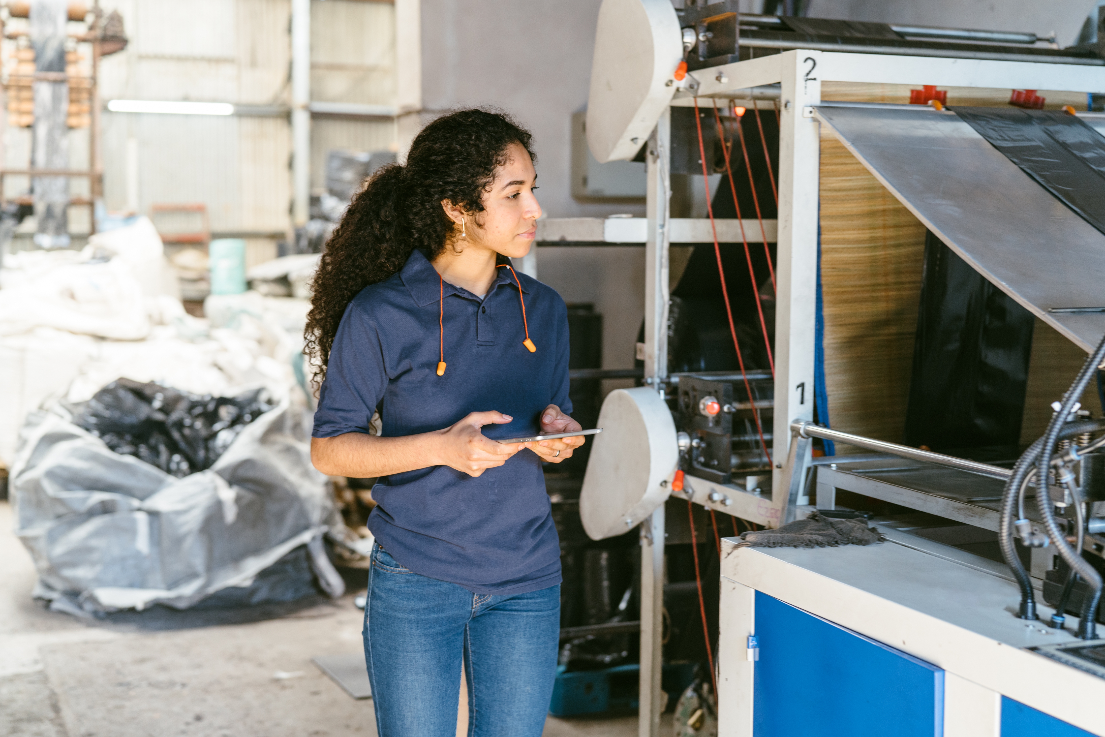 Person operating industrial machinery, focused and attentive, wearing casual attire with a tablet in hand. Surroundings feature large bags and equipment