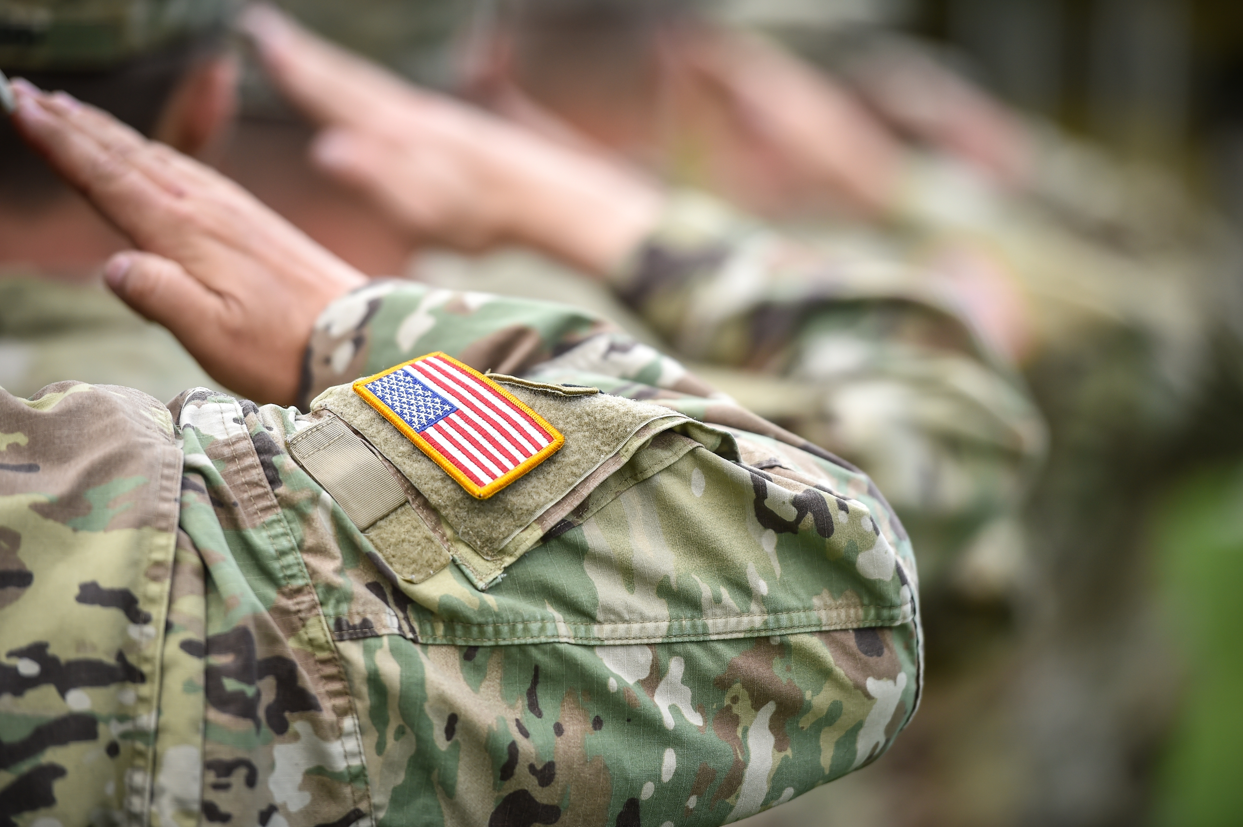 Soldiers in camouflage uniforms salute, displaying American flag patches, symbolizing service and dedication