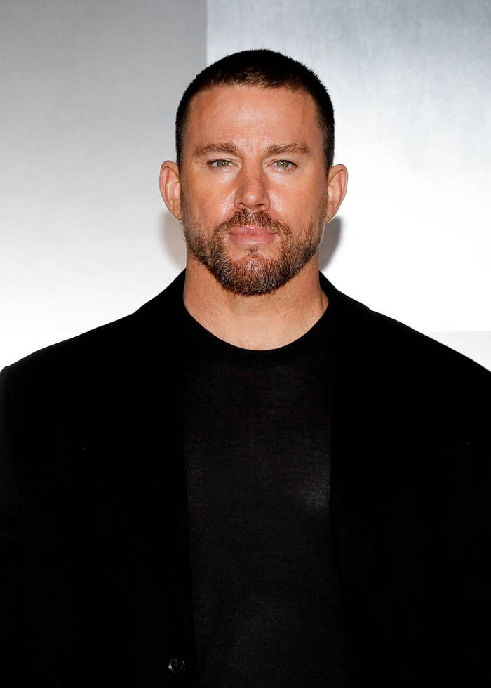 Channing Tatum with short hair and beard in a black suit jacket over a black shirt, posing against a plain background