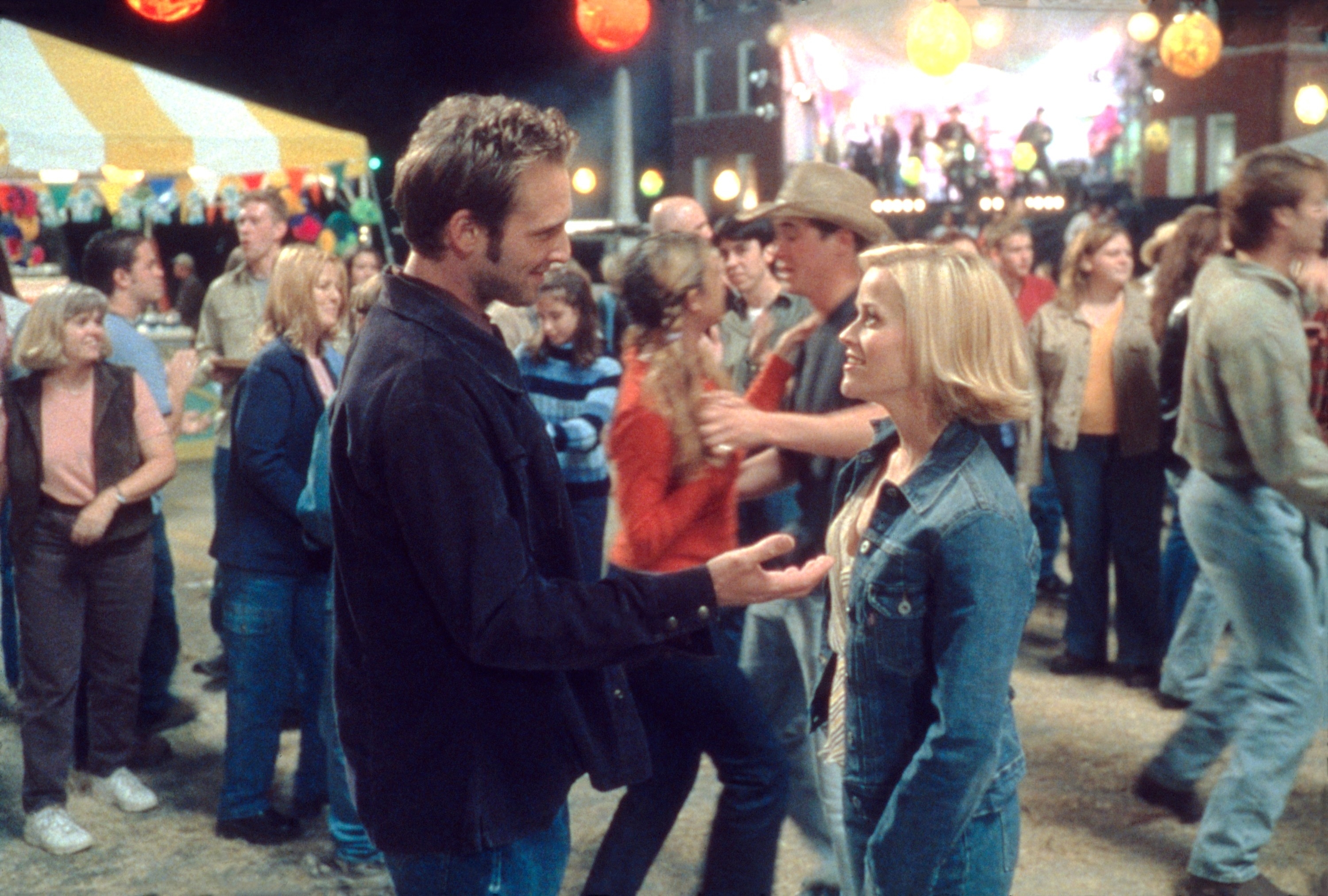Two people converse at an outdoor festival with a crowd in the background, suggesting a scene from a TV show or movie