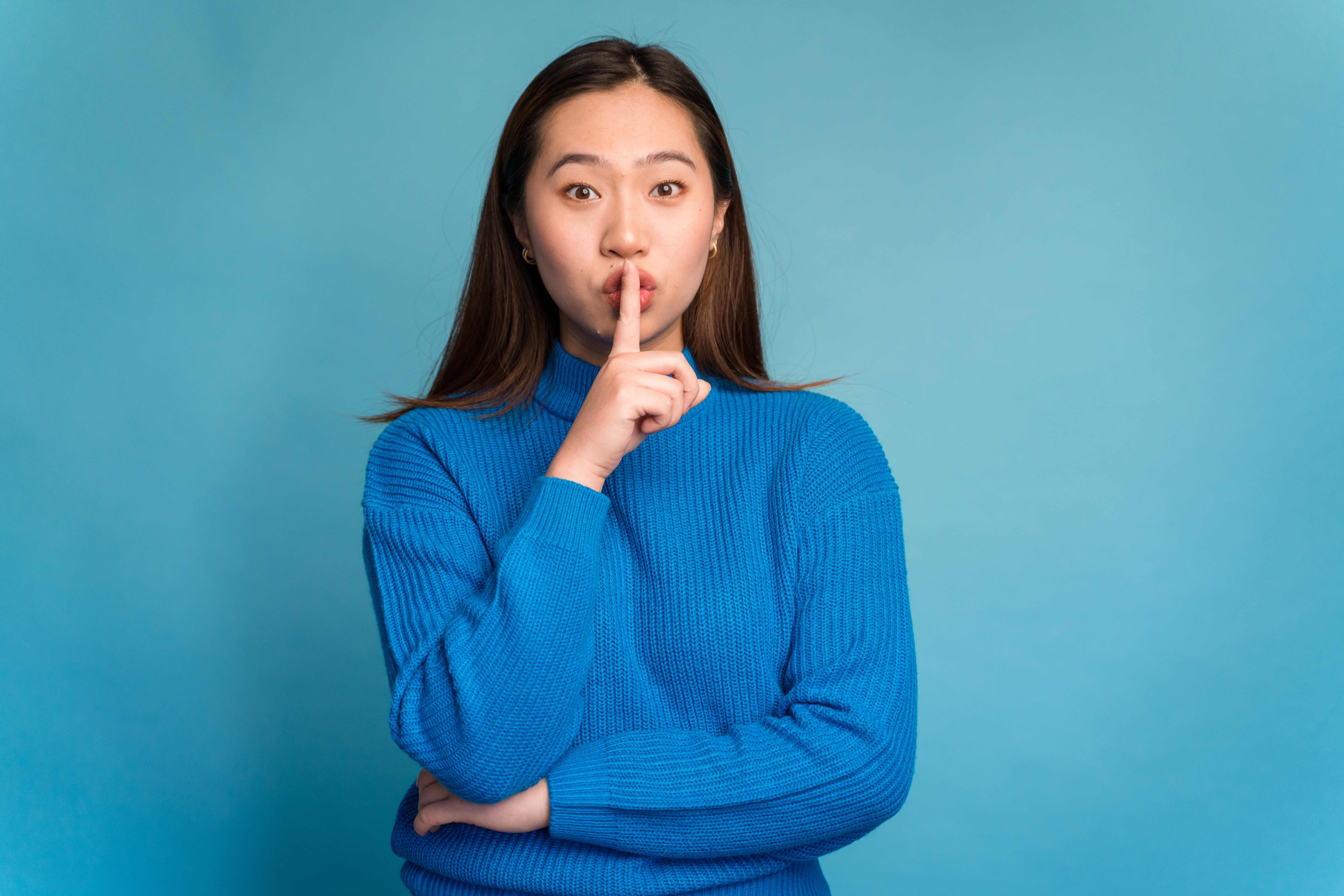 Person in a ribbed sweater holds finger to lips, gesturing for silence, against a plain backdrop