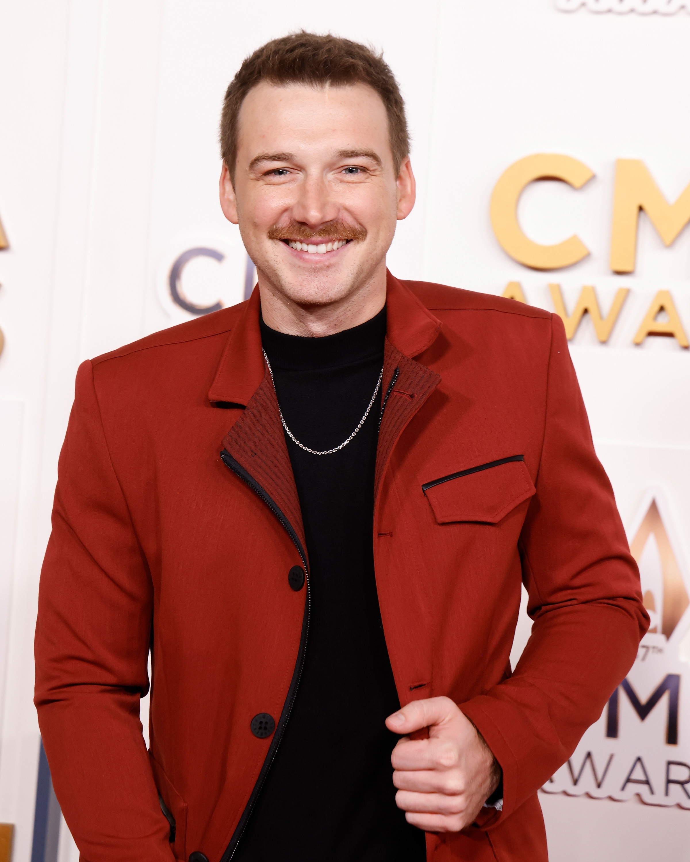 Smiling person on red carpet, wearing a tailored jacket with a black shirt and a chain necklace, at the CMA Awards