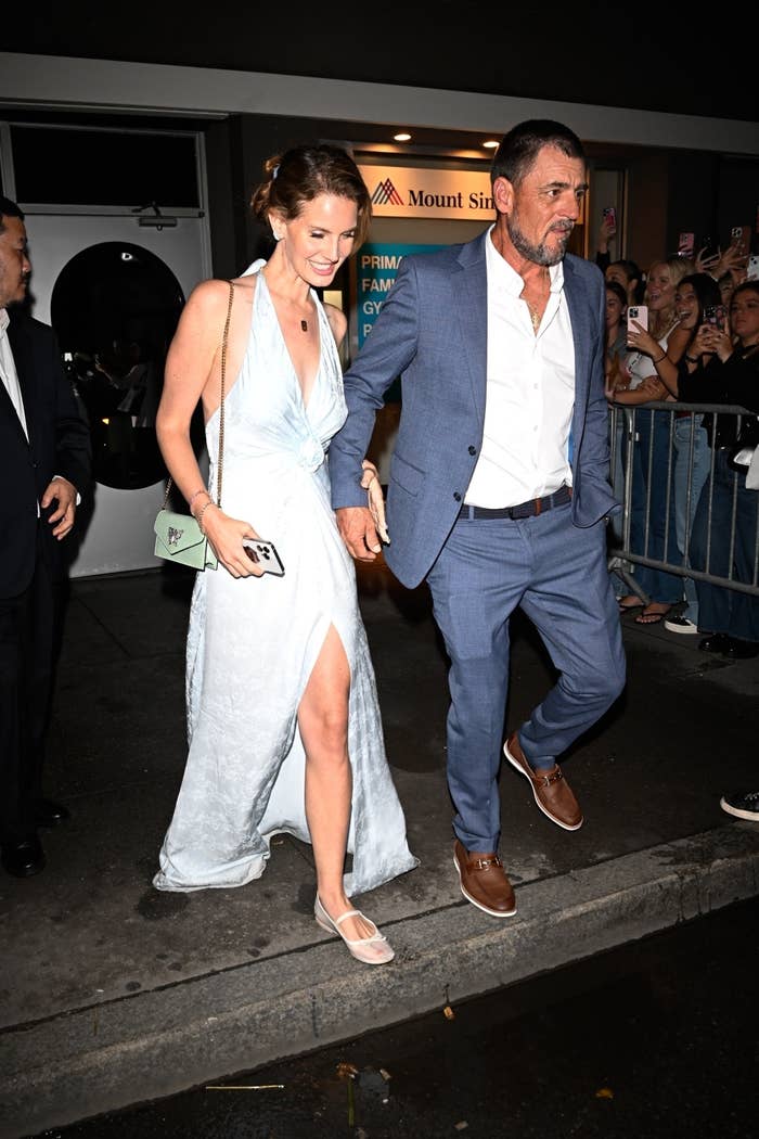 A woman in a light dress and a man in a suit are walking hand in hand outside, surrounded by onlookers