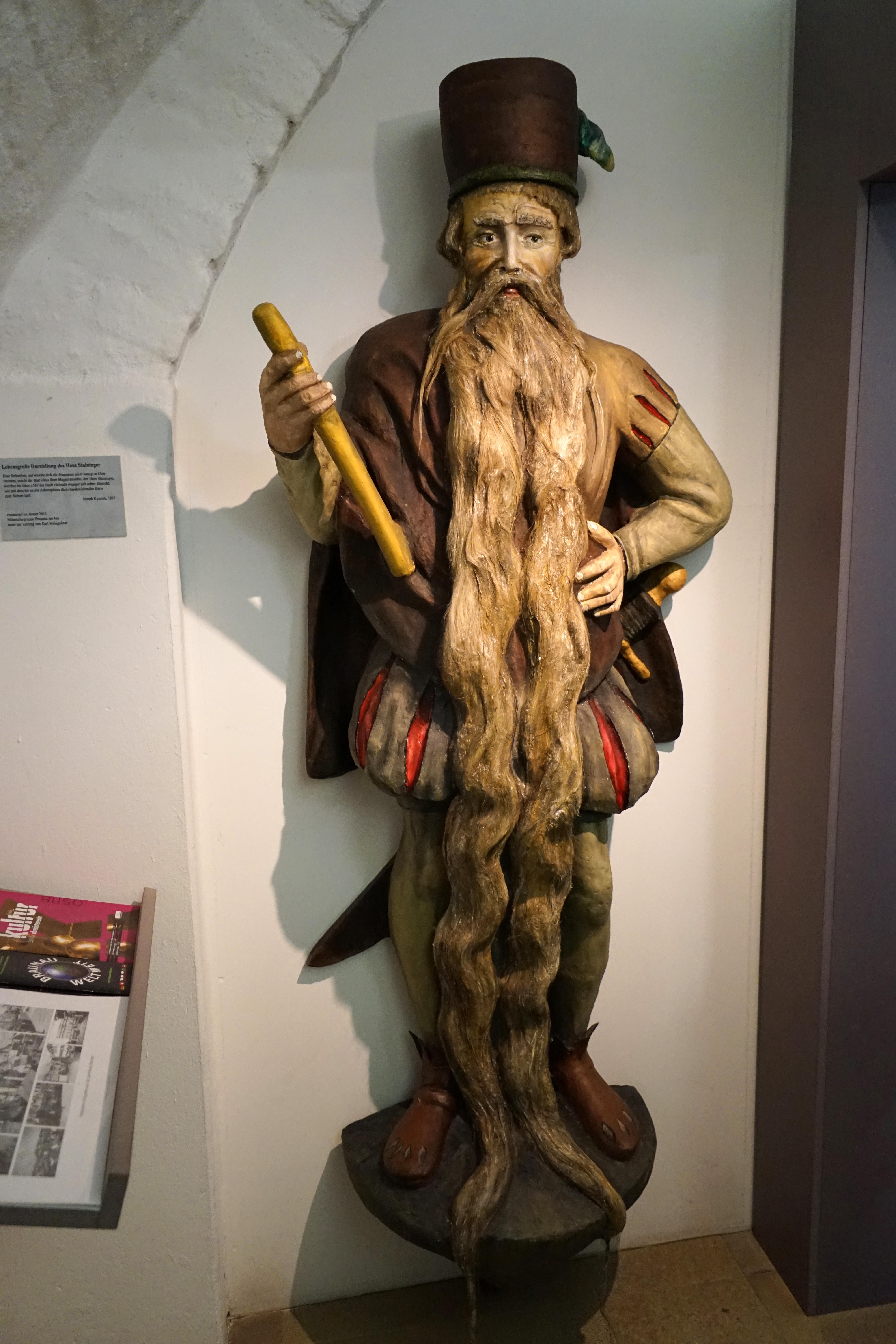 Statue of a bearded man holding scrolls, with his long beard intricately styled, displayed in a museum setting