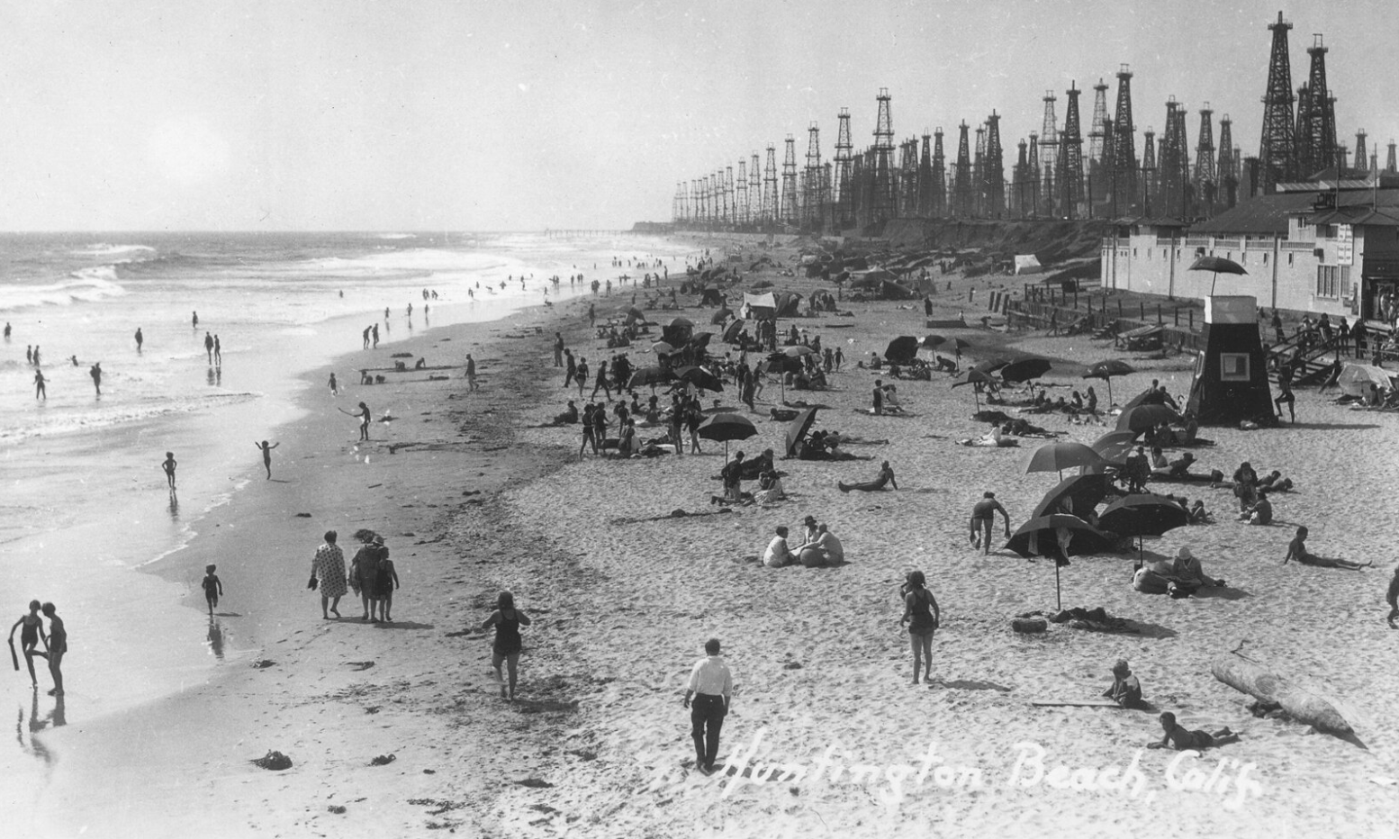 People enjoy a busy Huntington Beach, California, in a vintage photo with many oil derricks visible in the background