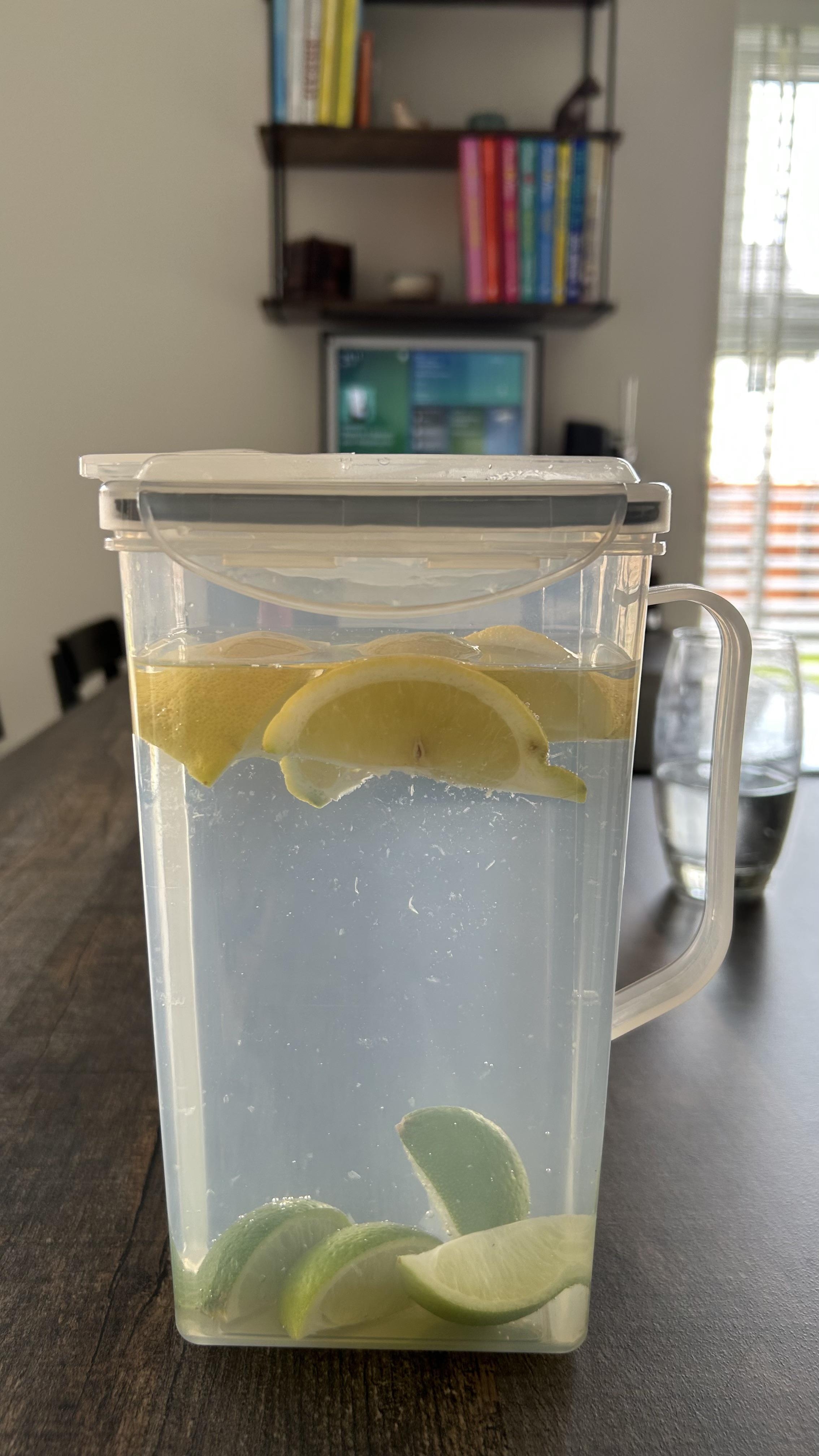 A clear pitcher of water with lemon and lime slices on a dark table in a home setting with a shelf and TV in the background