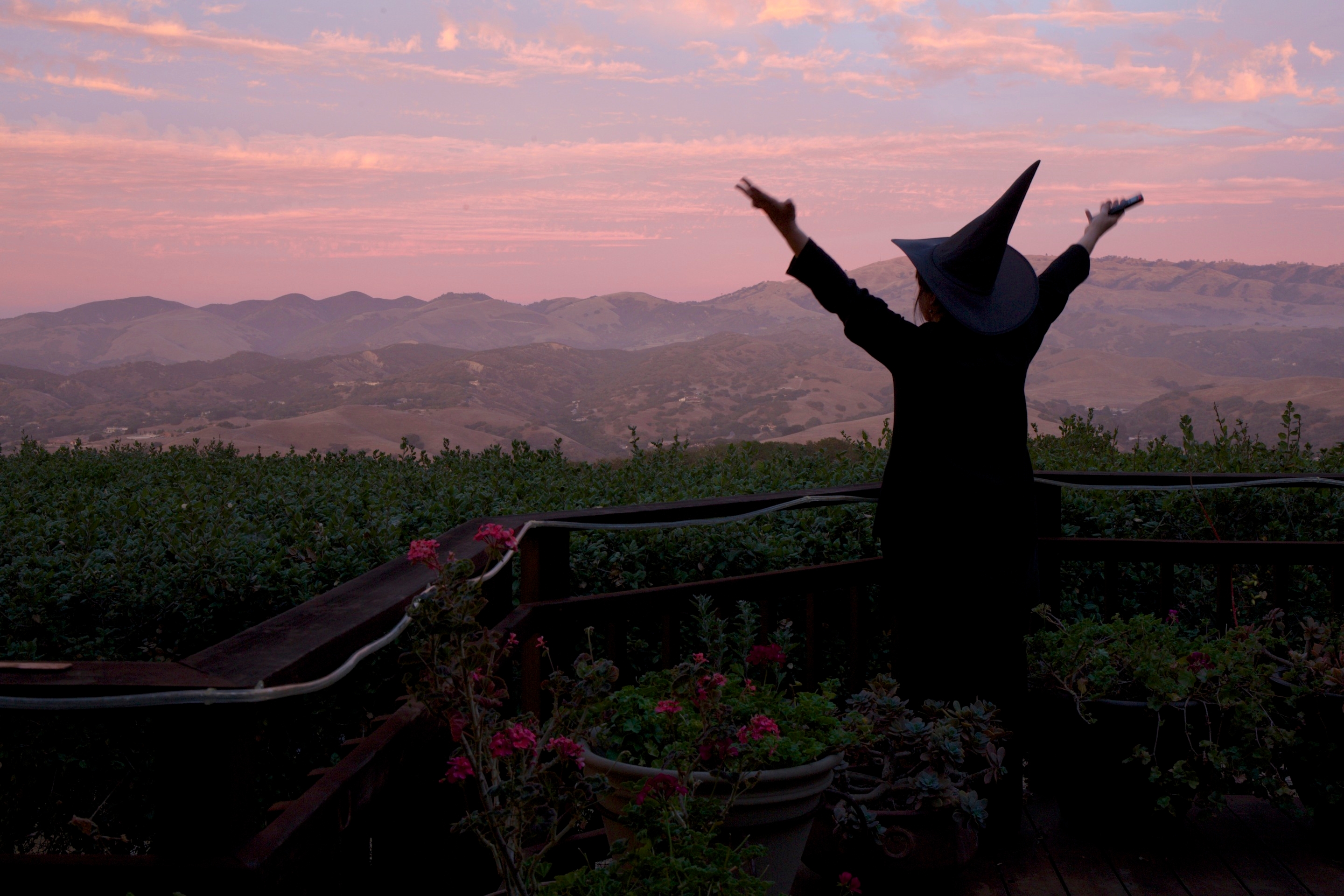 Person in a witch costume joyfully raising arms at sunset on a deck overlooking rolling hills