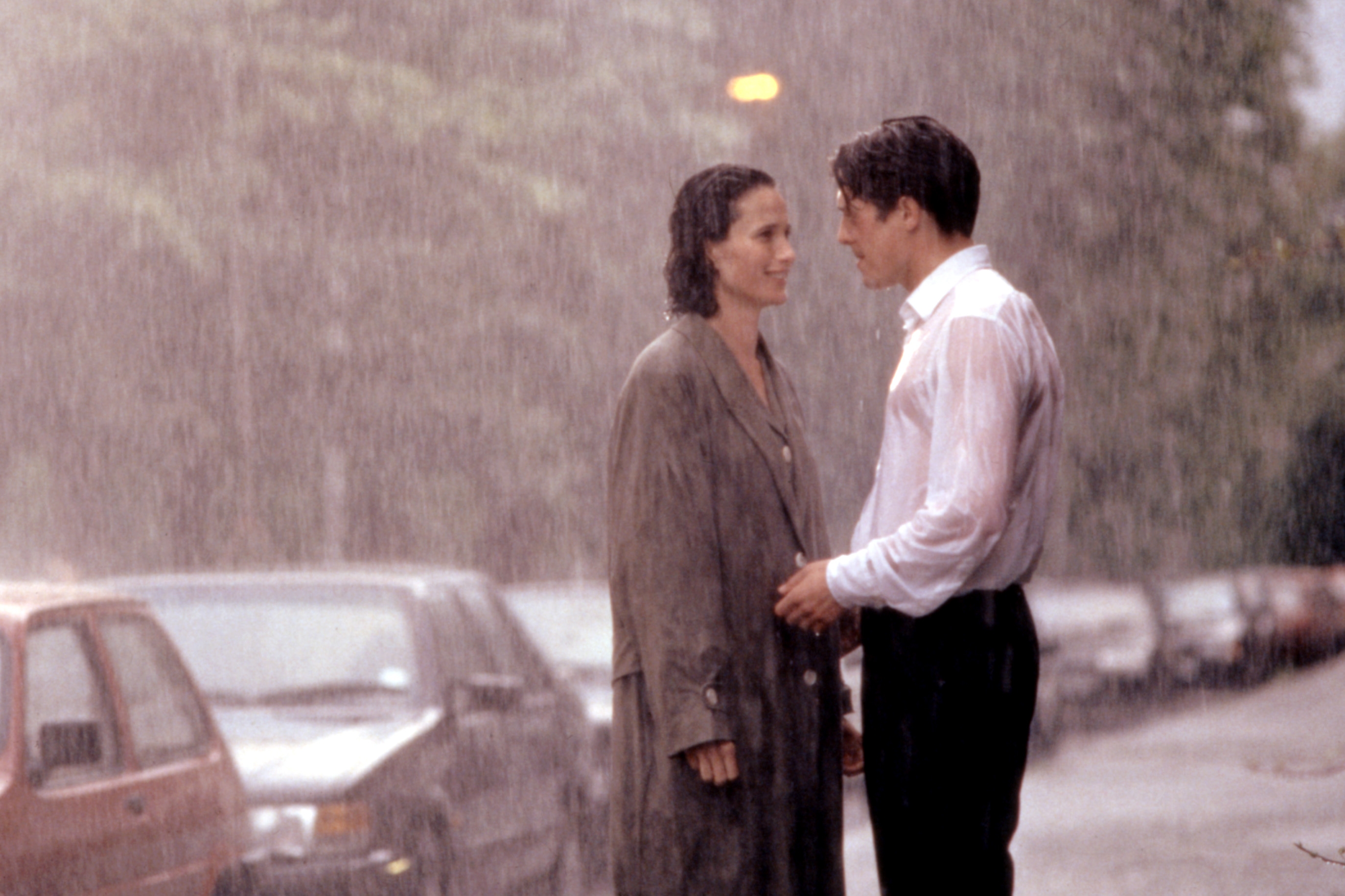 Two people in a romantic scene, standing close in the rain on a street. Both wear professional attire, engaged in an emotional moment