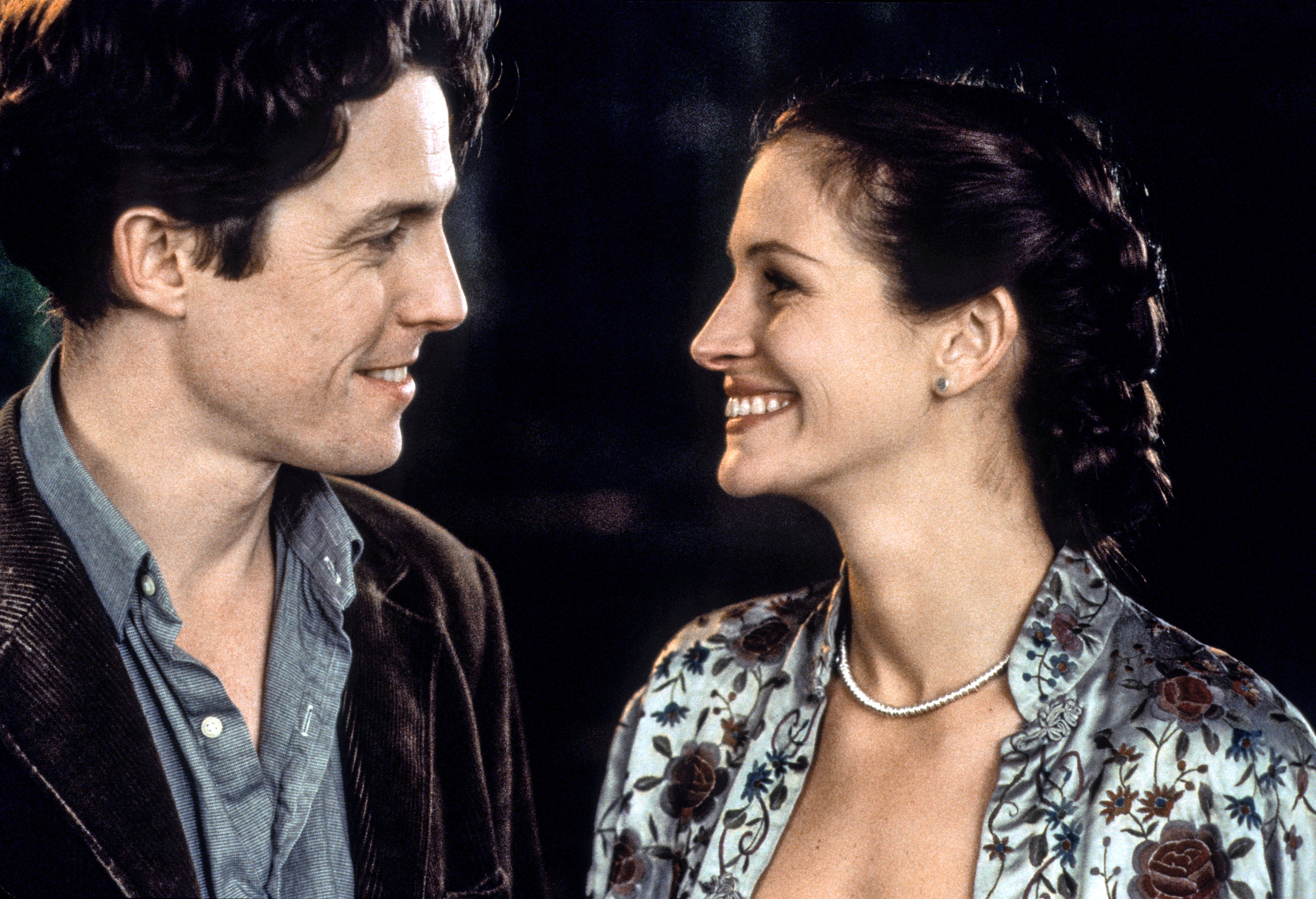 Two actors in a romantic onscreen moment, smiling at each other. The woman wears a floral blouse and pearl necklace while the man wears a casual jacket