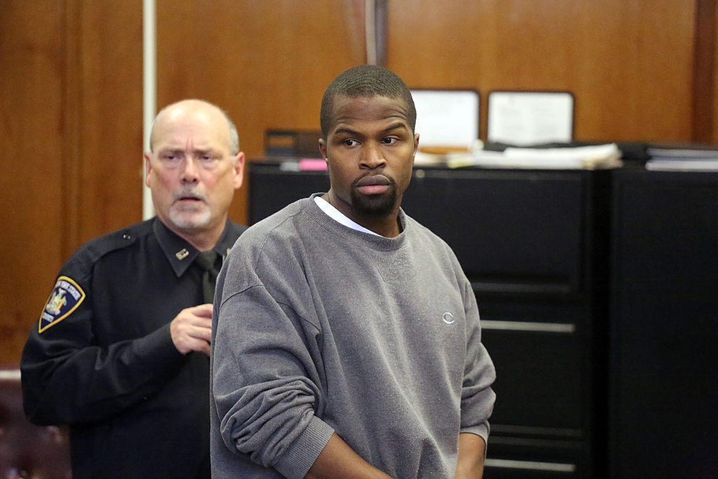 A man in a courtroom is wearing a casual sweatshirt, standing with an officer nearby
