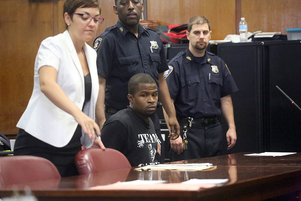 A person is seated at a courtroom table, surrounded by three officers and a person in a white blazer