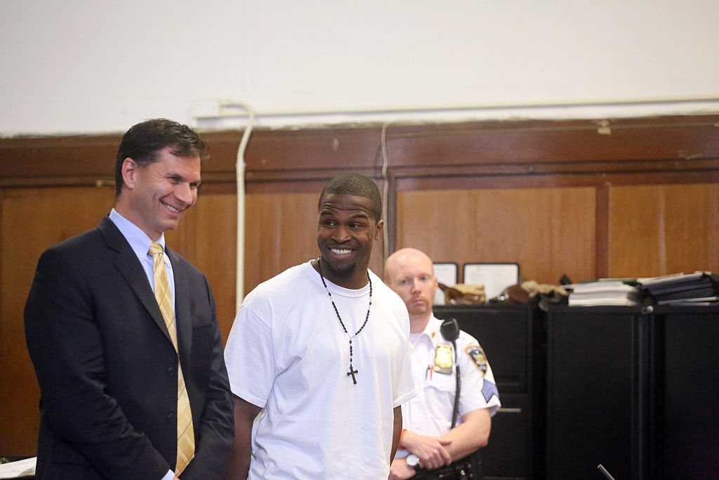 Two men standing in a courtroom; one is dressed in a suit while the other wears a casual outfit with a visible cross necklace. A security officer is nearby