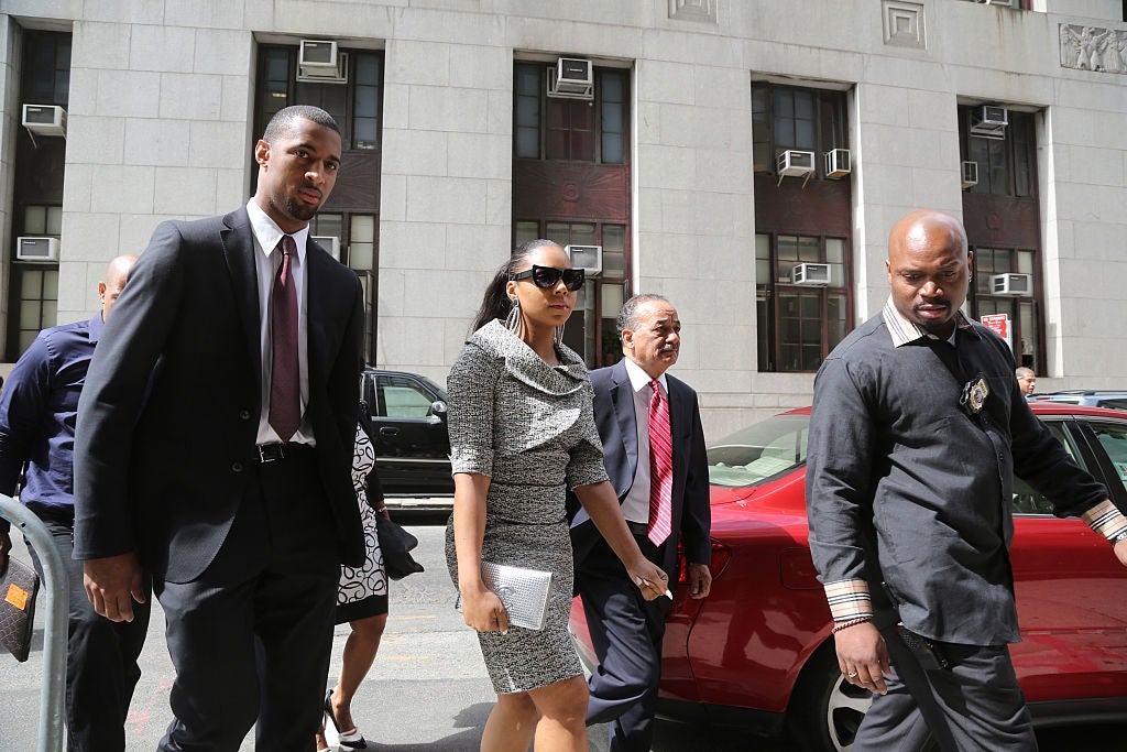 A woman in a stylish dress walks with a group of people outside a building, suggesting a high-profile event or legal appearance