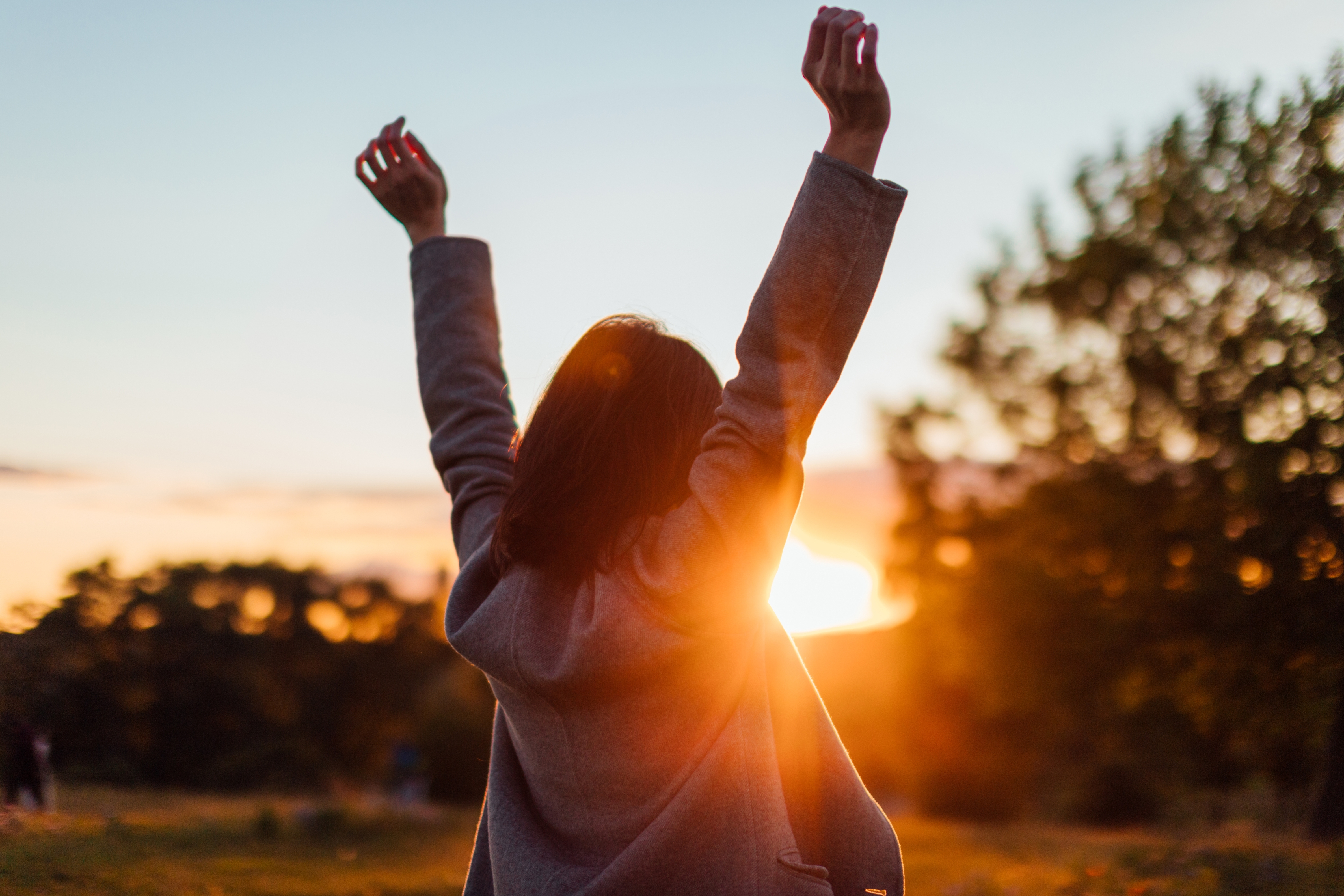 Person outdoors with arms raised, facing sunrise. Silhouette against a scenic background of trees and sky. Peaceful and uplifting moment