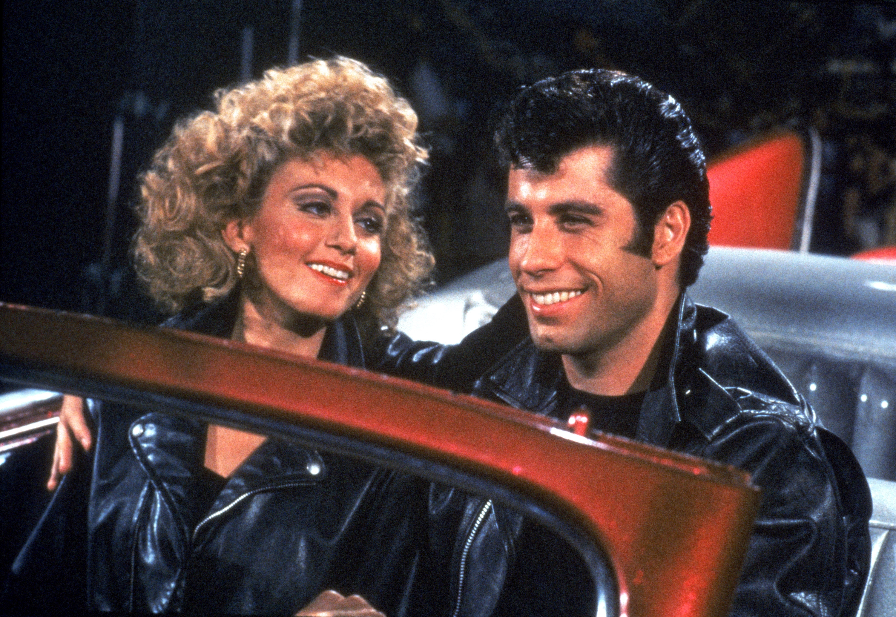 Two actors in leather jackets smiling while sitting in a car, evoking a classic movie scene atmosphere