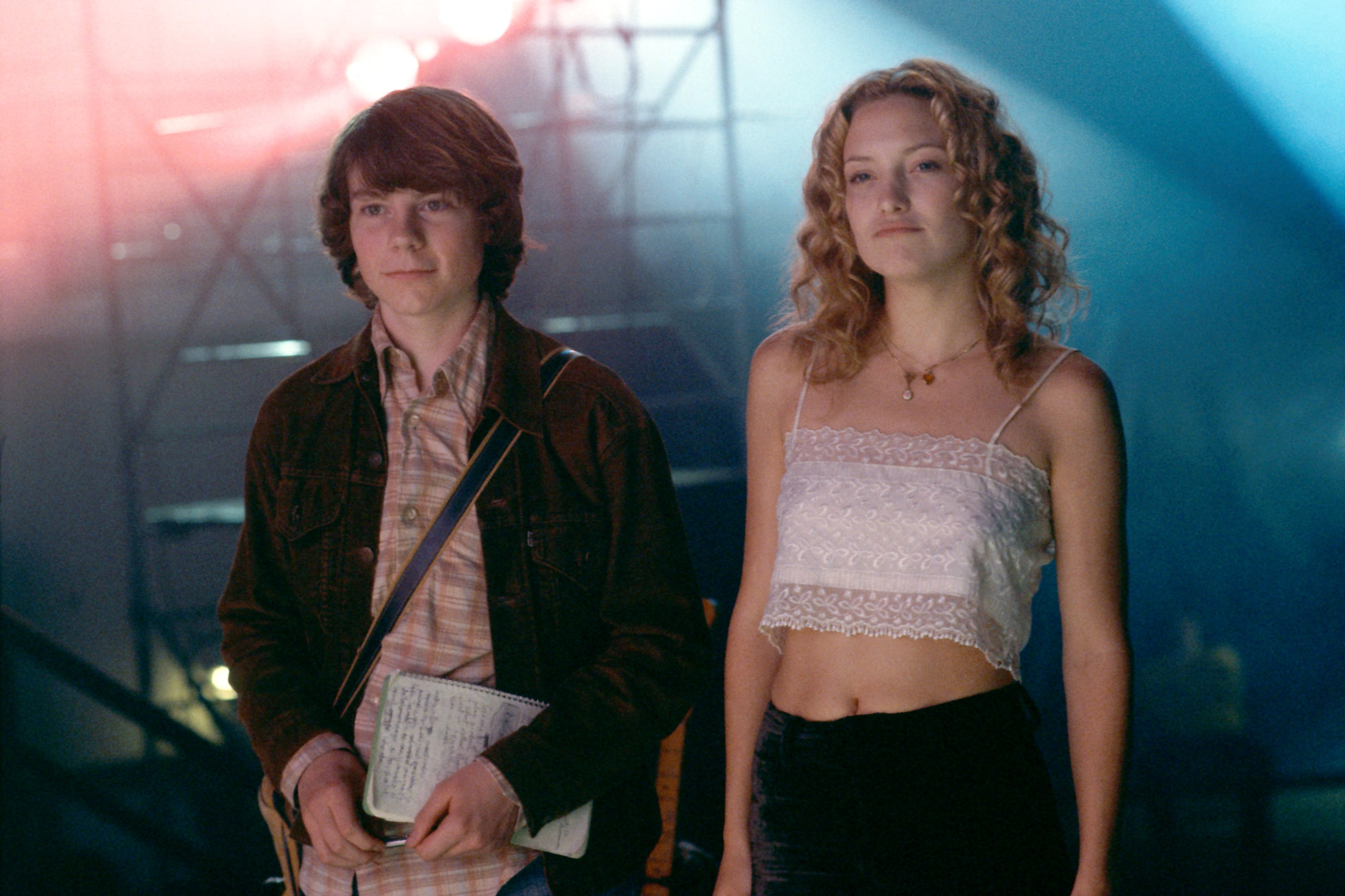 Two people standing together, one with a notebook and wearing casual 70s-style attire; the other in a lace top and dark pants, featured in a film scene