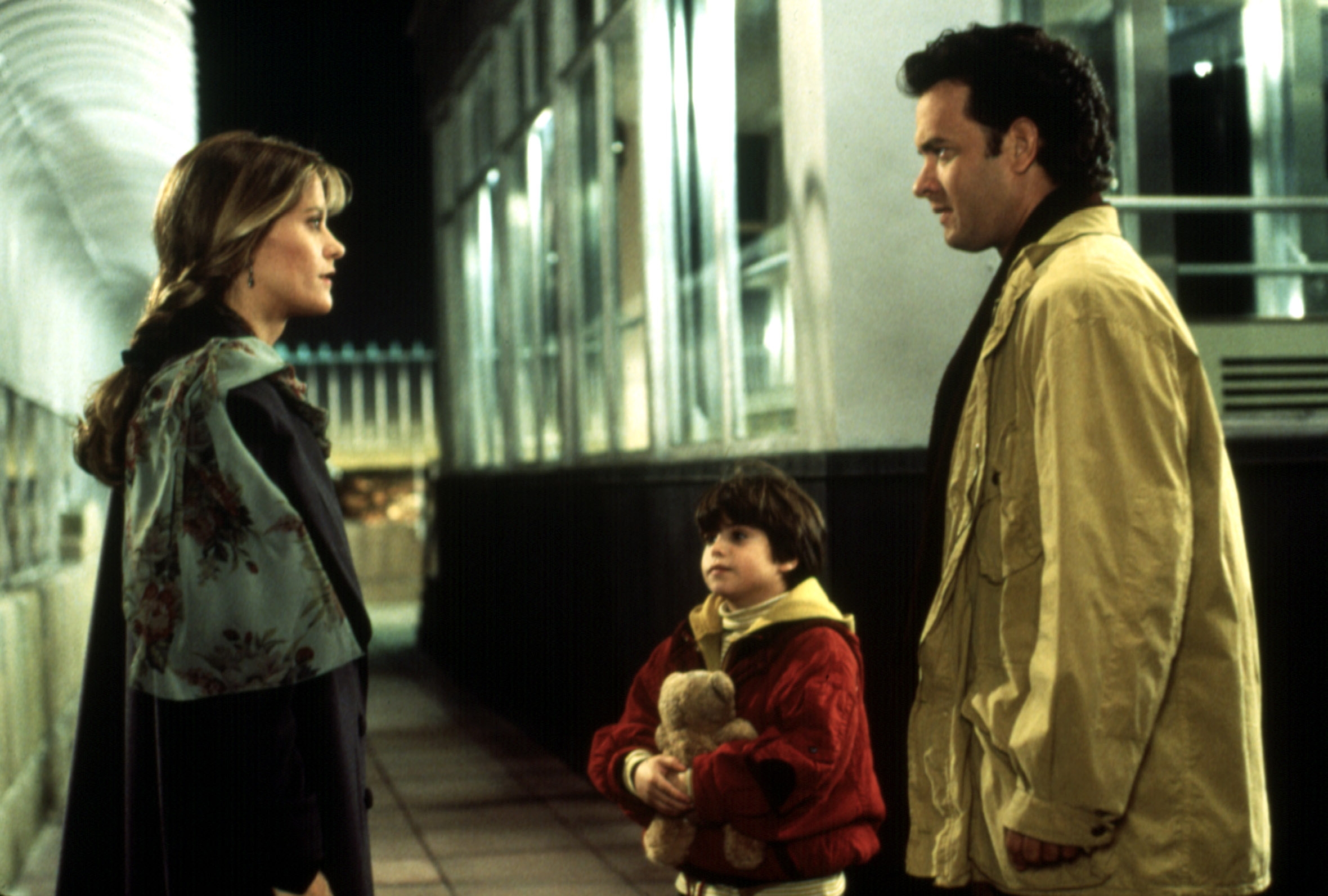 Three people stand on a city observation deck. A woman and a man face each other, with a child holding a teddy bear between them