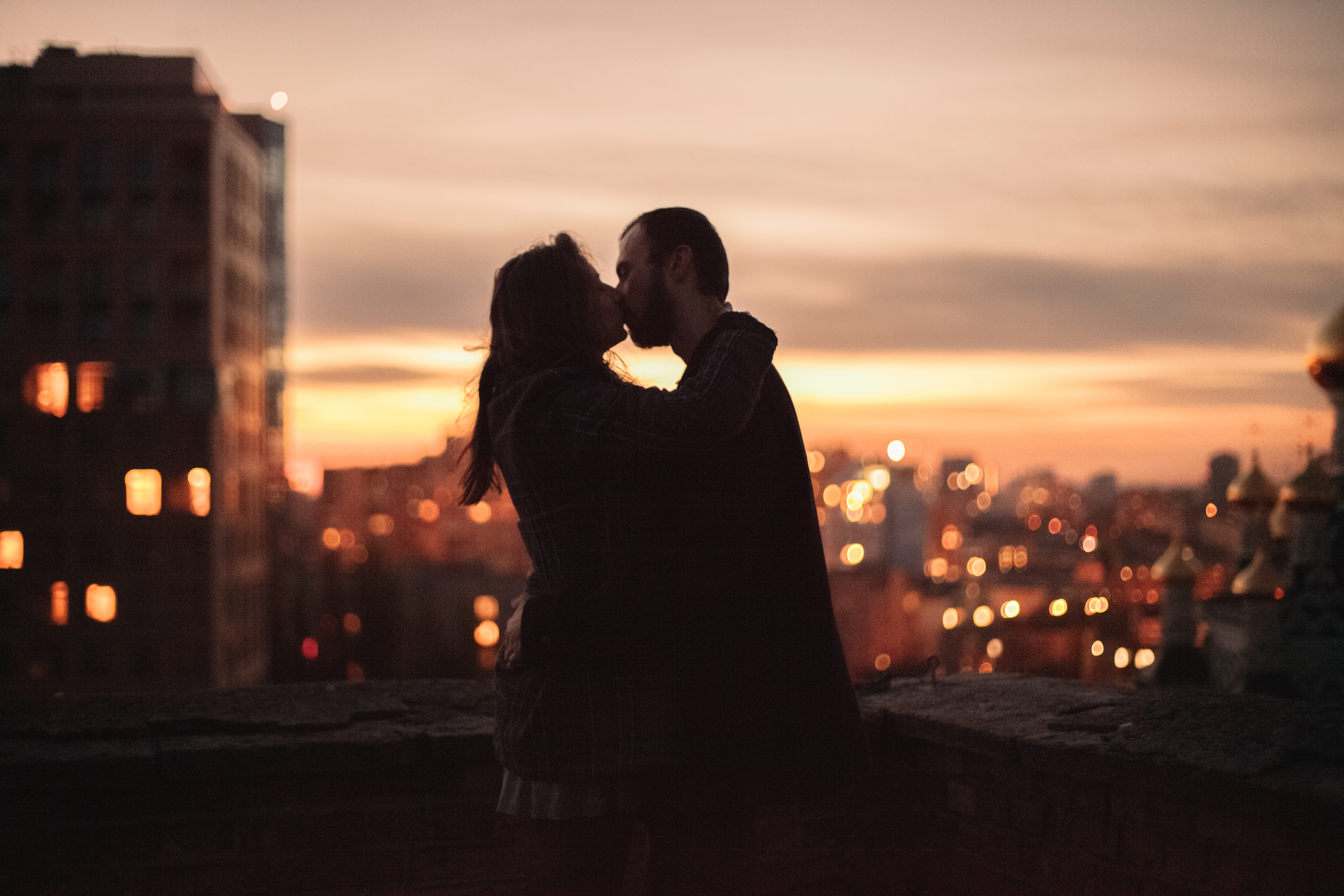 A silhouette of a couple embracing at dusk on a city rooftop with glowing lights in the background