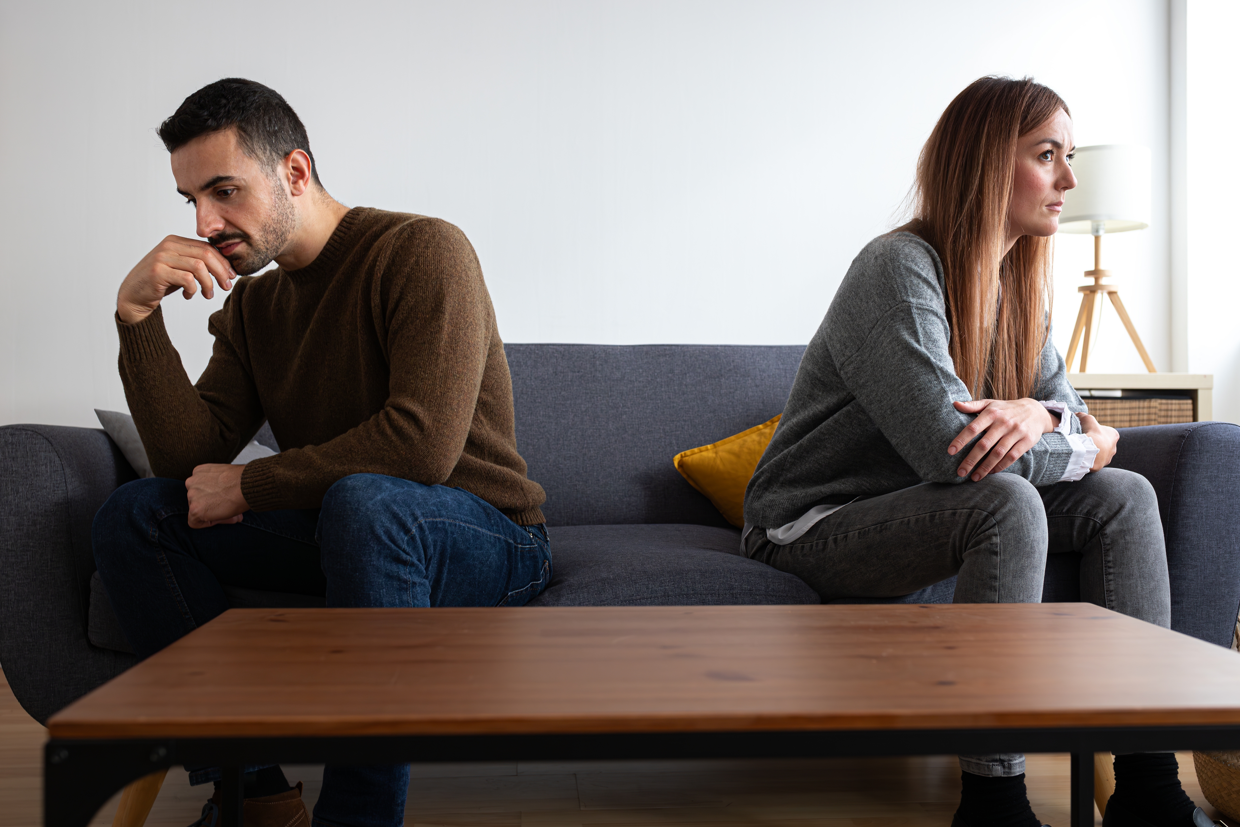 A couple sits on a sofa, looking away from each other, appearing deep in thought or possibly upset