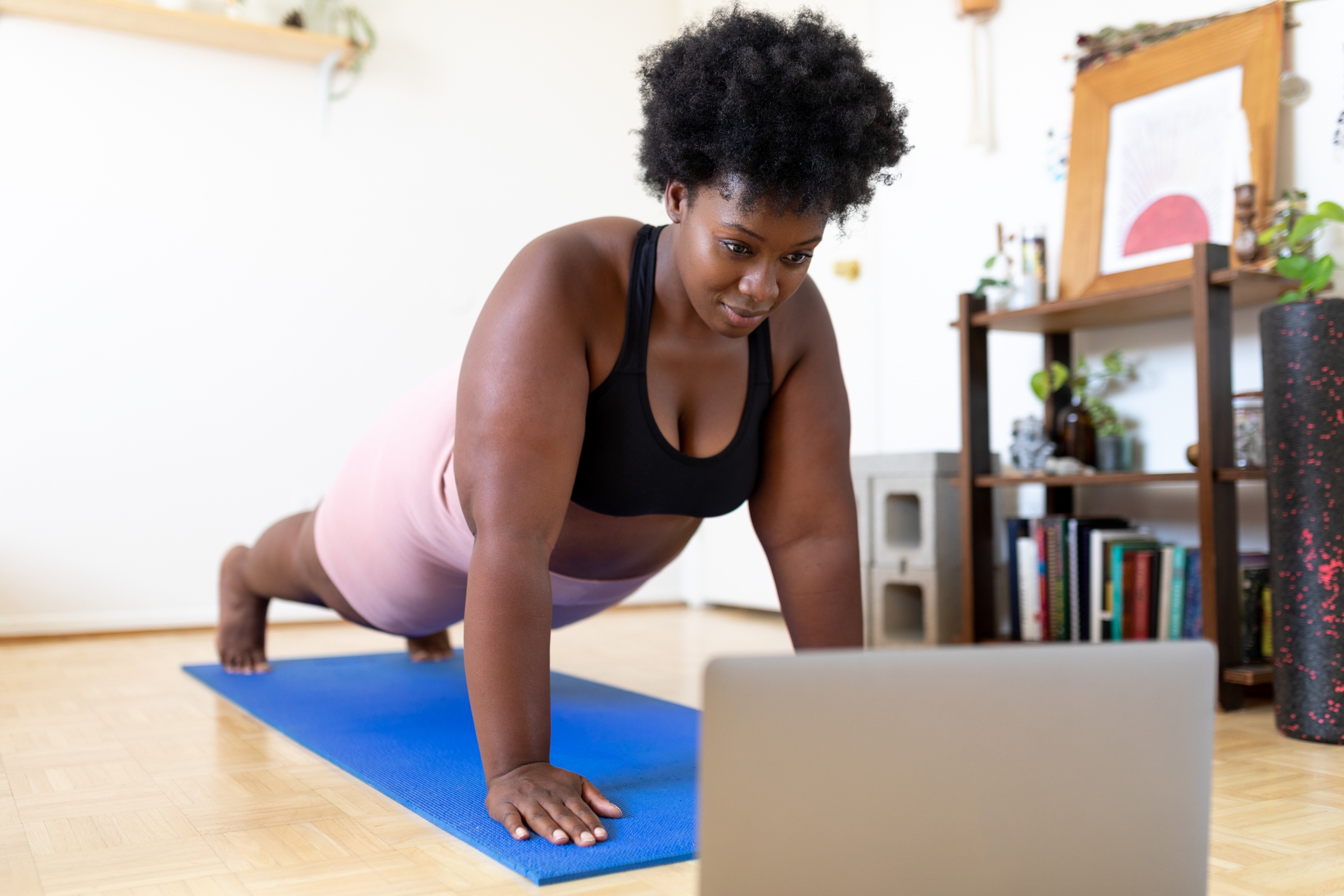 Person som gjør yogaplank på en matte, ser på en bærbar datamaskin for veiledning i et koselig, hjemmemiljø med planter og bøker