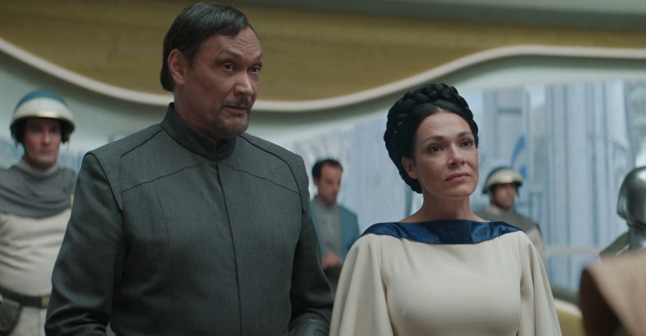 Two people in formal, futuristic attire discuss in a sci-fi setting, with others in the background wearing uniforms and helmets