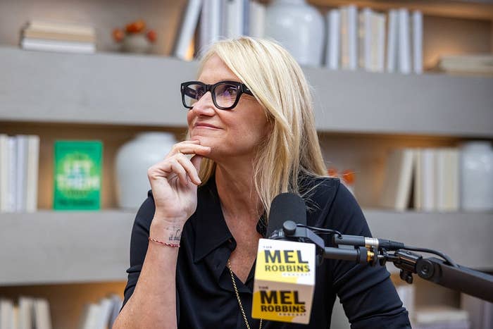 Person with glasses, smiling, sits at a table with a microphone labeled "Mel Robbins." Background has shelves with books and decorative items