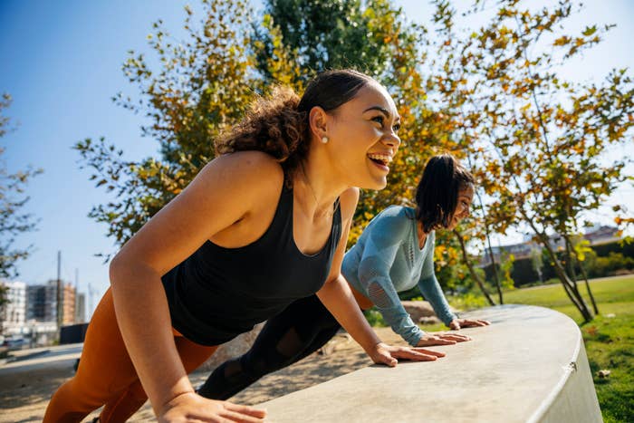 Two women doing push-ups outdoors, smiling and enjoying a sunny day in the park, surrounded by trees and buildings in the background