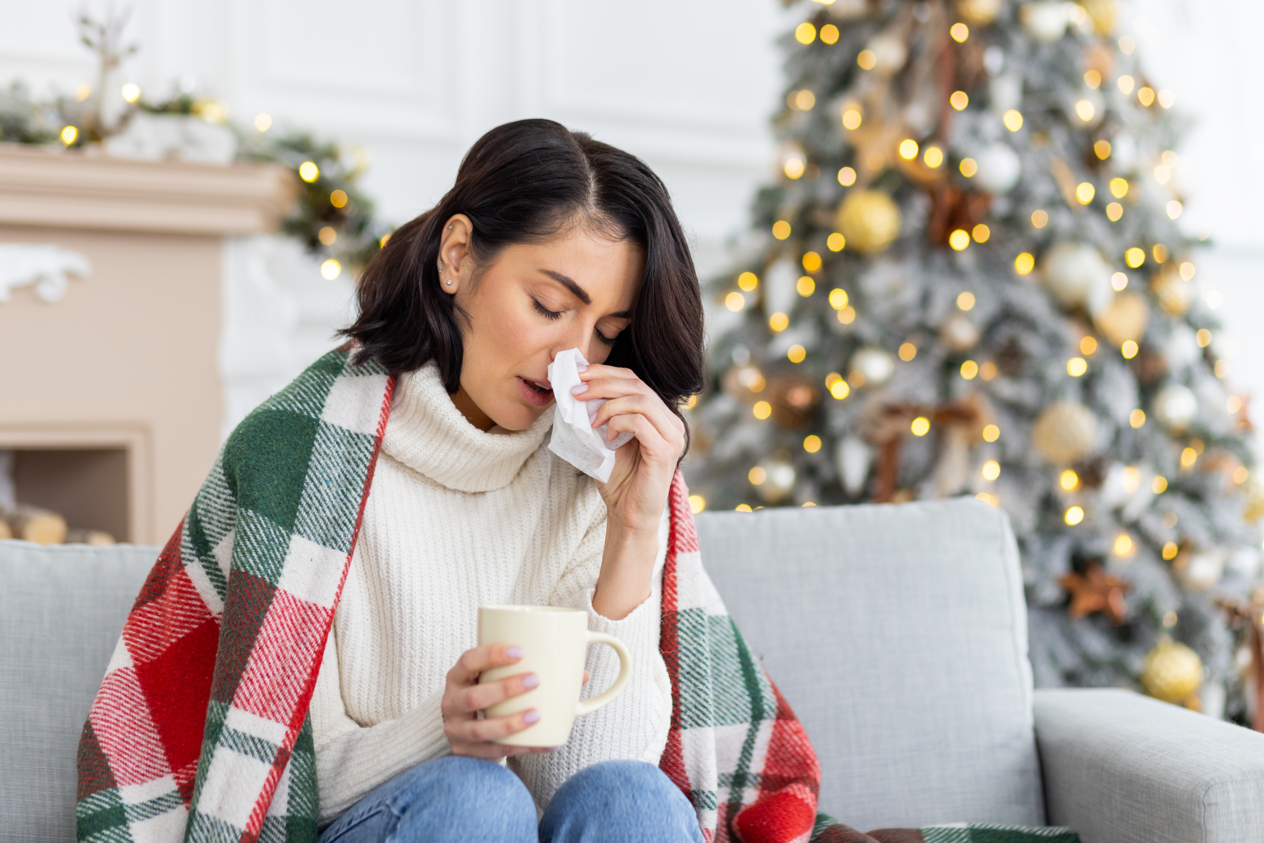 Woman on couch with tissue and mug, wrapped in a plaid blanket, Christmas tree in background