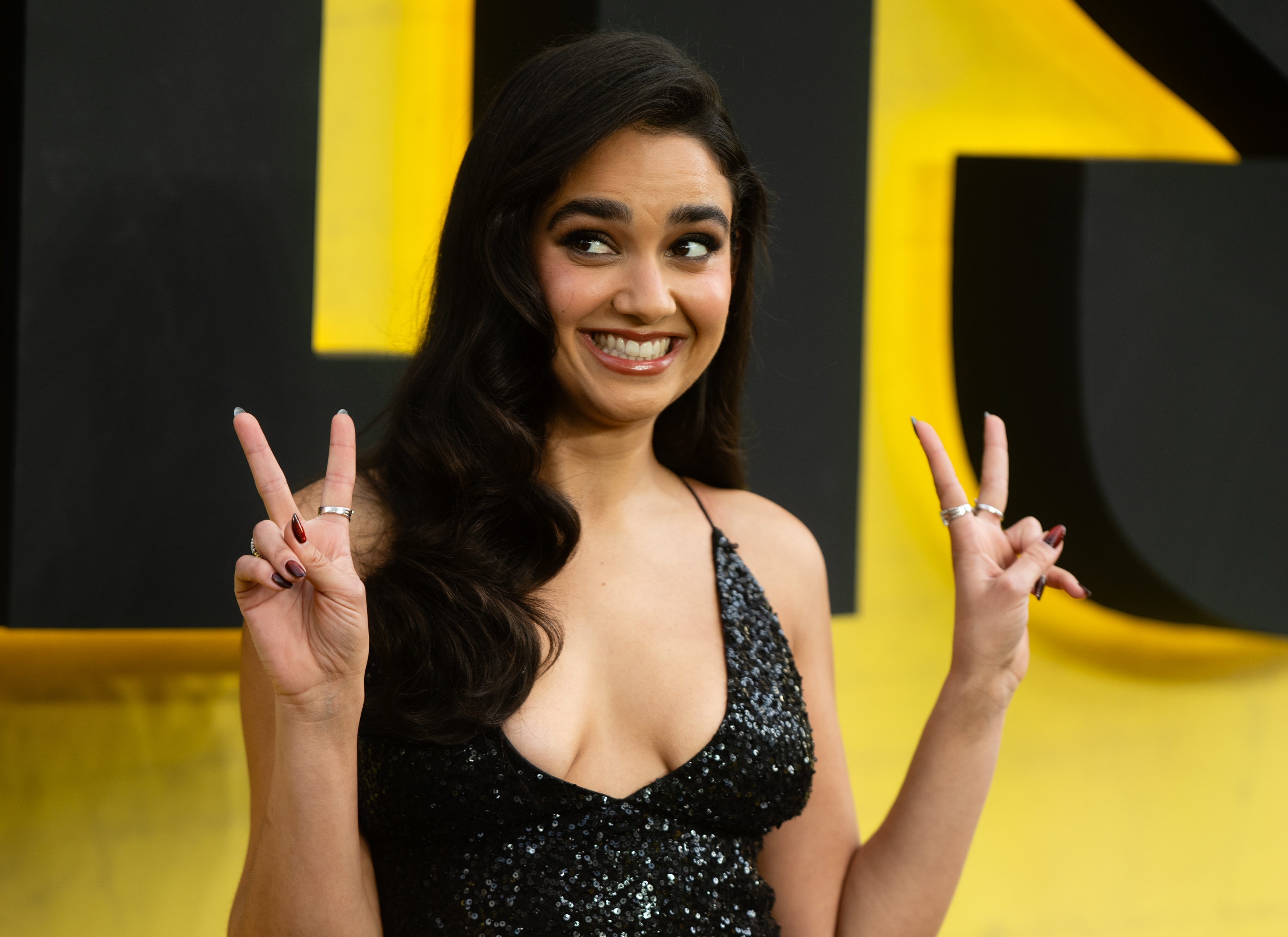 Person on red carpet in a sparkling black gown, smiling with both hands showing peace signs