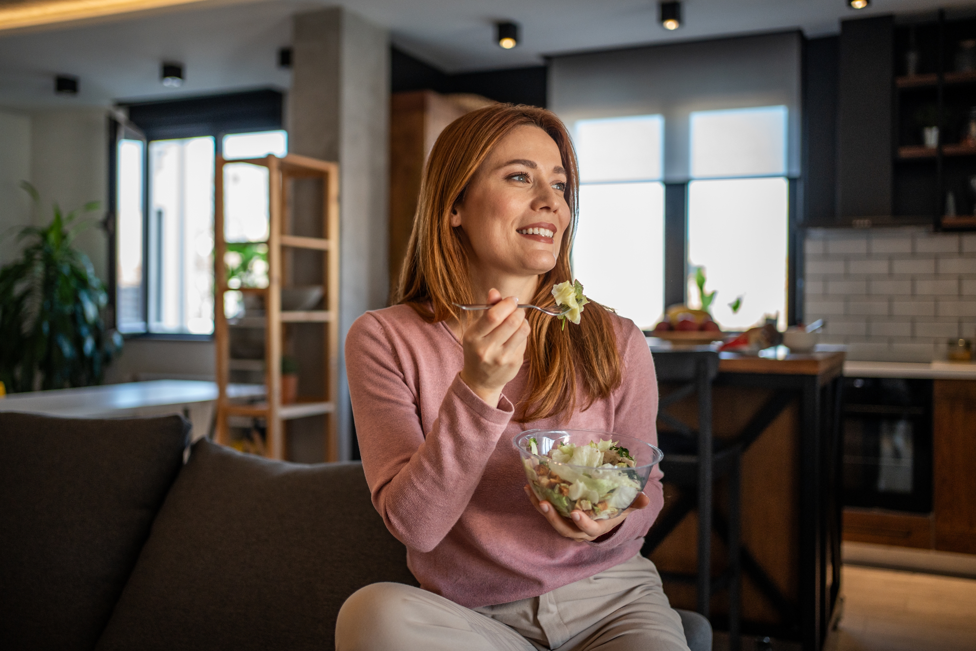 A person is seated on a sofa in a modern kitchen, smiling and eating a salad from a glass bowl