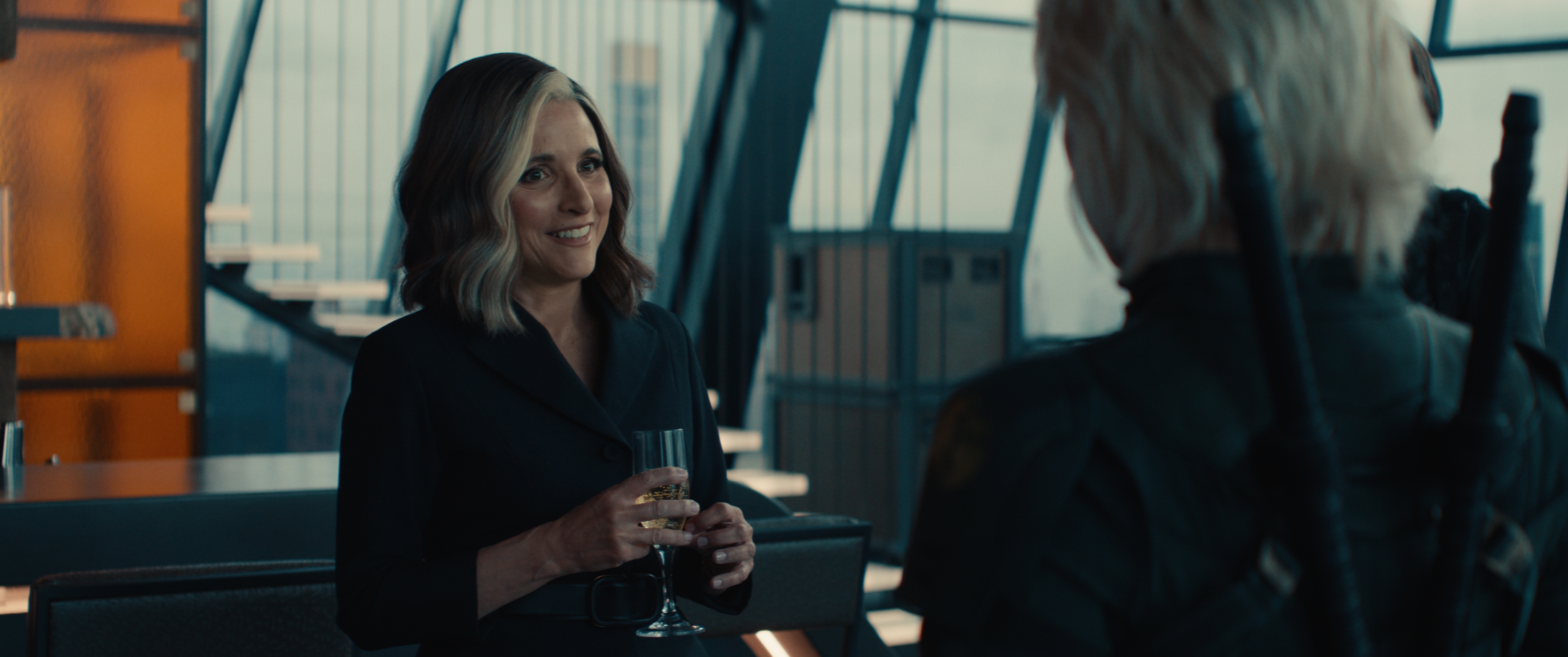 Woman in sleek, dark outfit holding a glass, talking to a person with short, light hair and sword sheaths, in a modern building