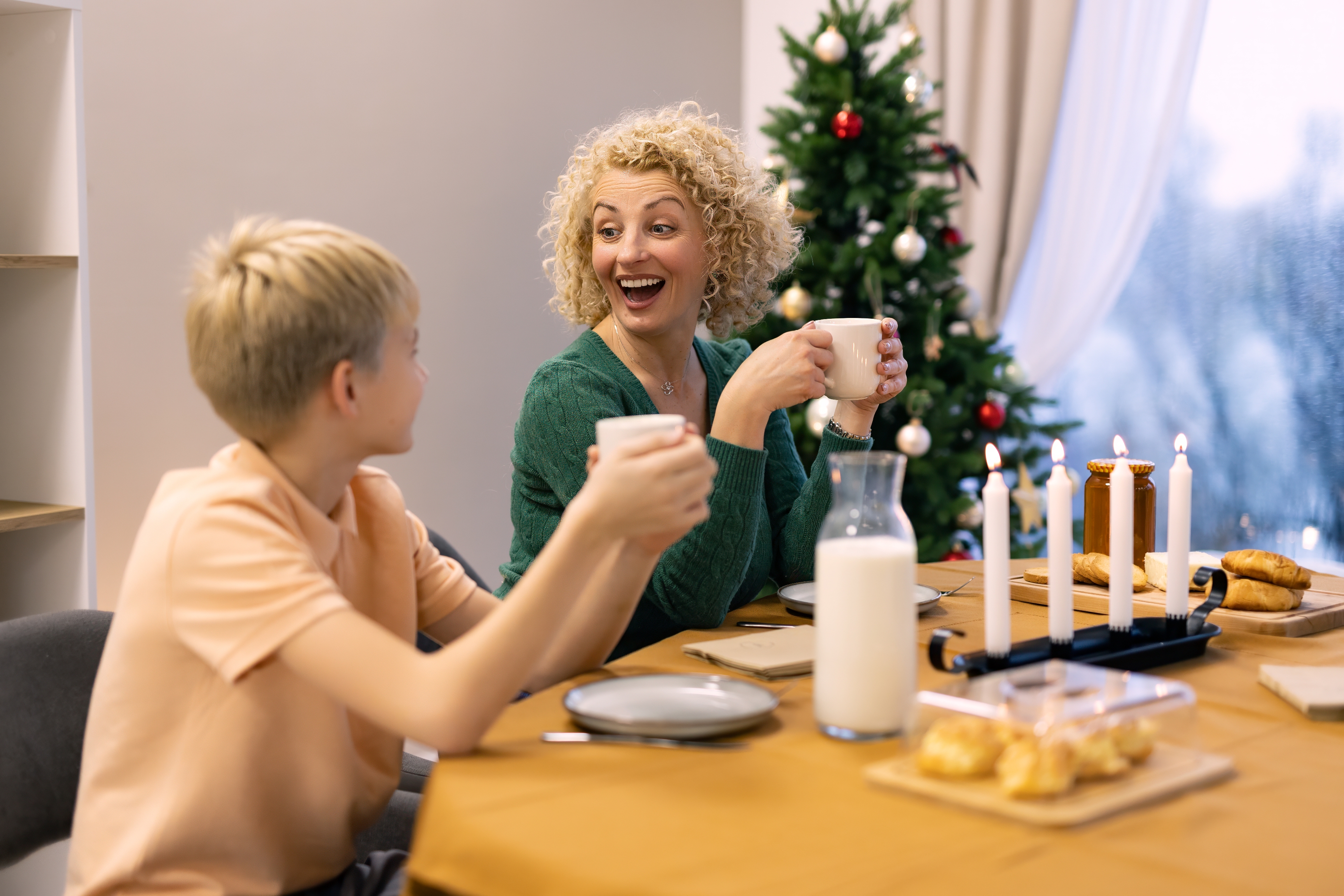 Woman and child smiling, holding mugs at a dining table with lit candles and a decorated Christmas tree in the background
