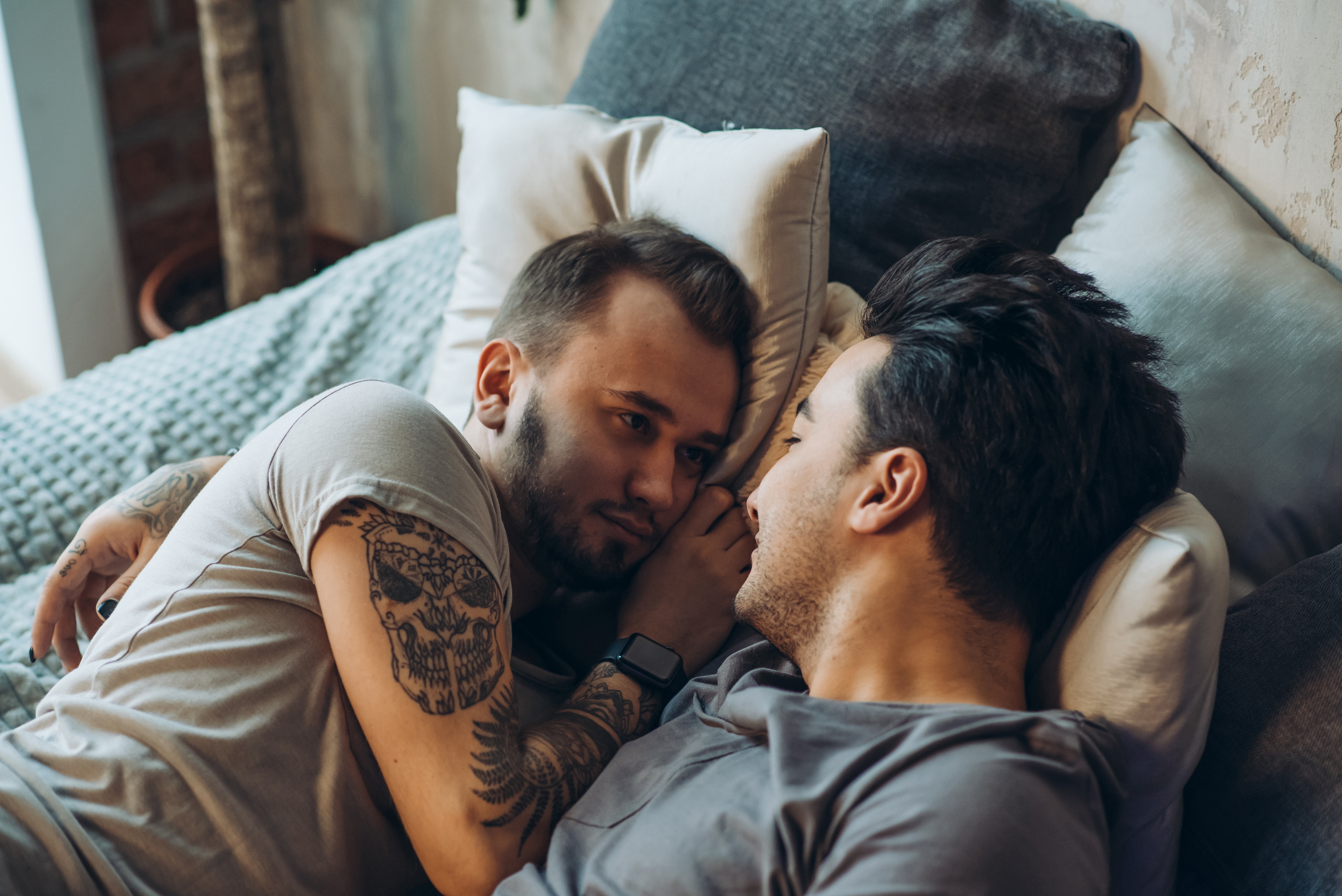 Two people lying in bed facing each other, one with a skull tattoo on their arm, sharing an intimate and tender moment