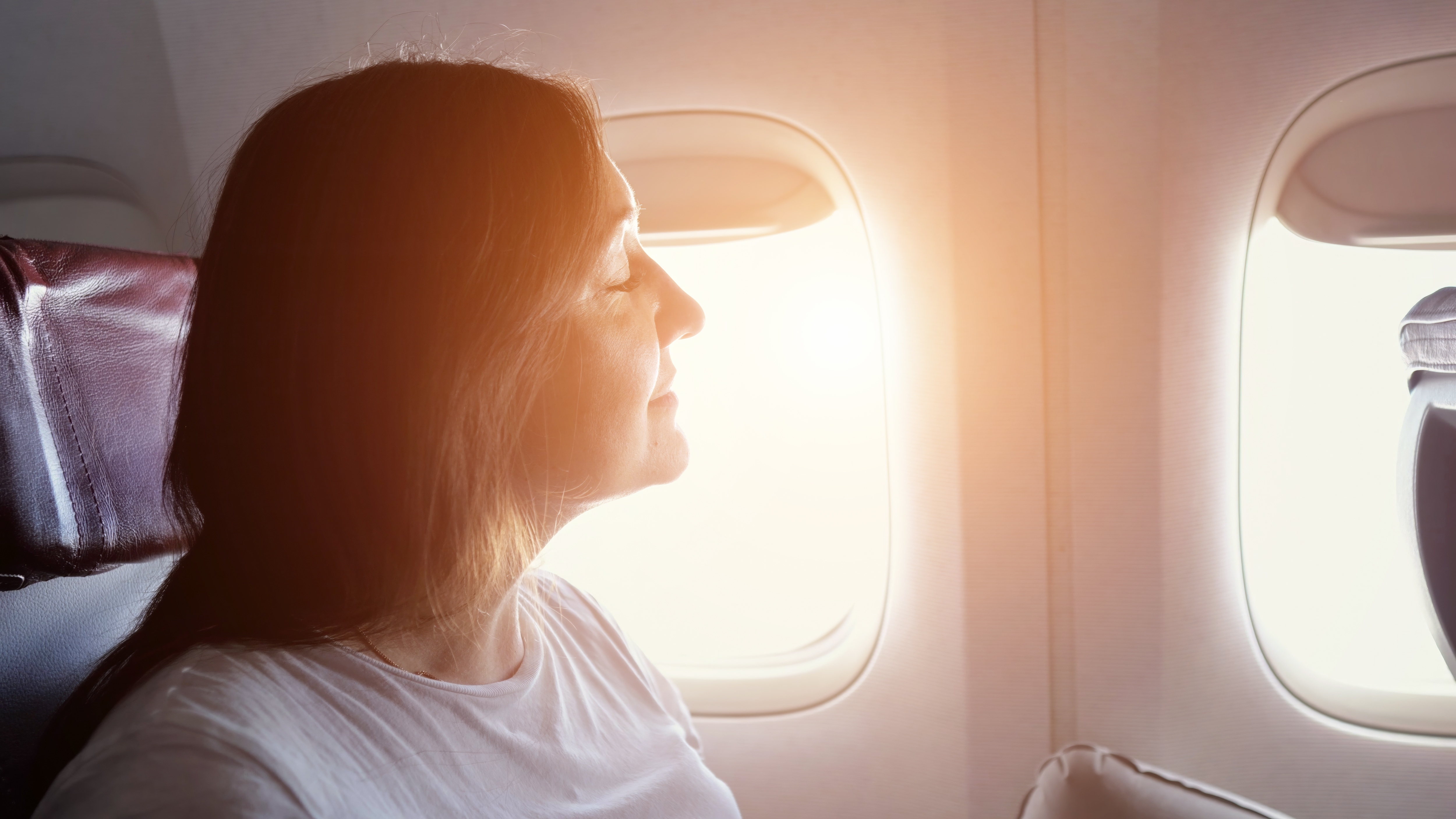 Person sitting by airplane window, eyes closed, relaxed expression, soft light from window illuminating face