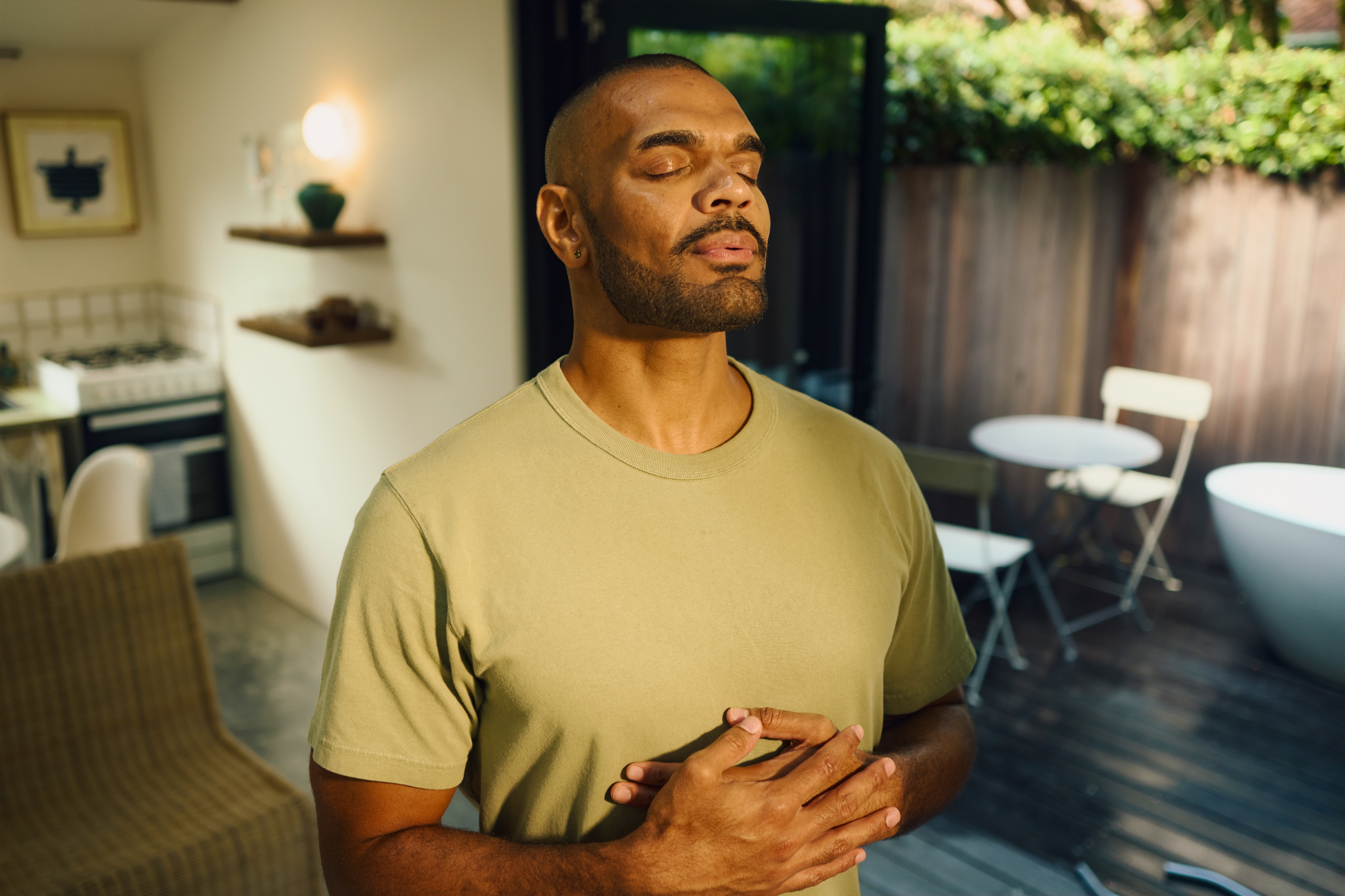 Person standing outdoors with eyes closed, hands on chest, appearing relaxed. Background shows patio furniture and a partially visible kitchen area
