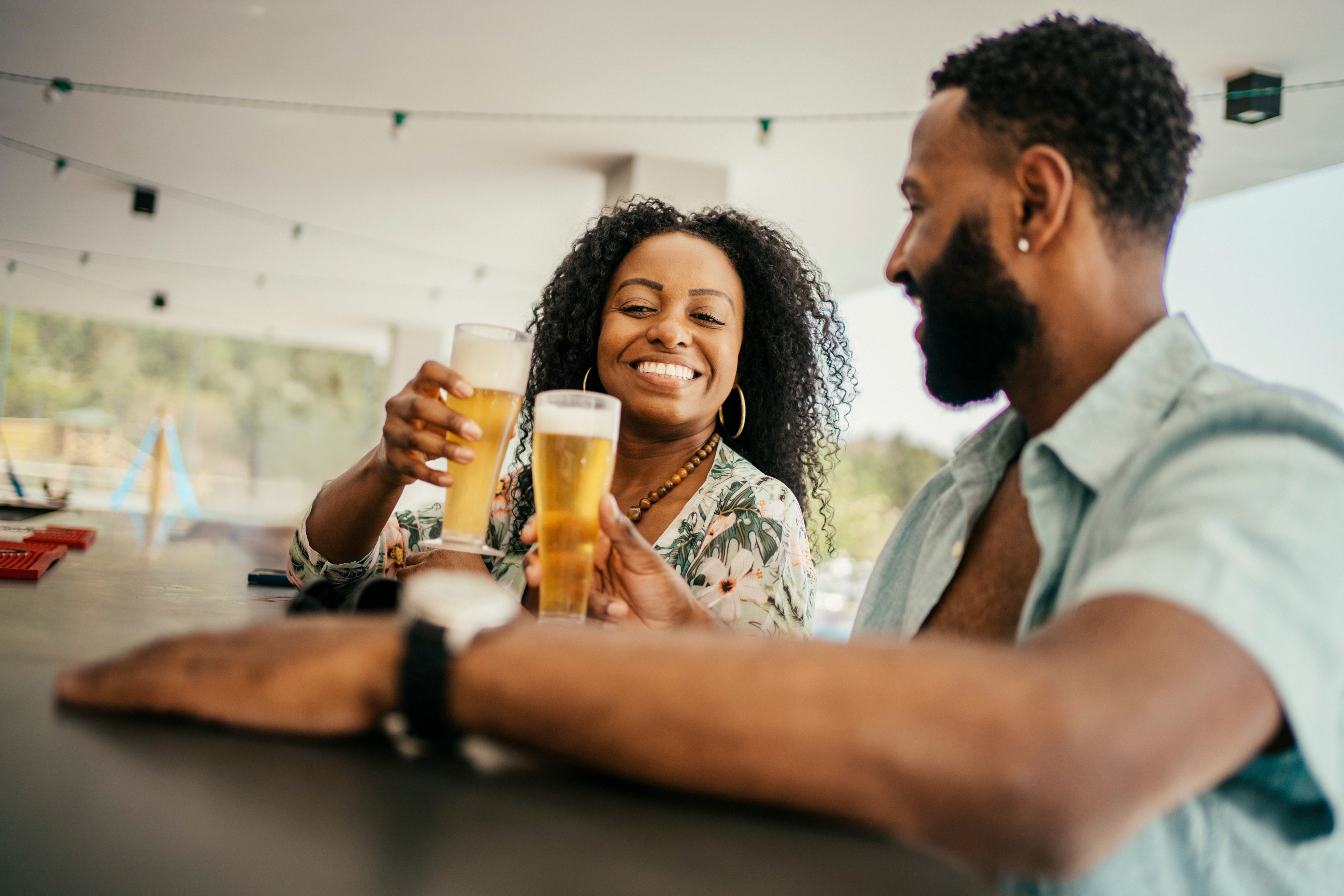 Two people smiling and clinking glasses of beer at a bar, enjoying a casual moment together