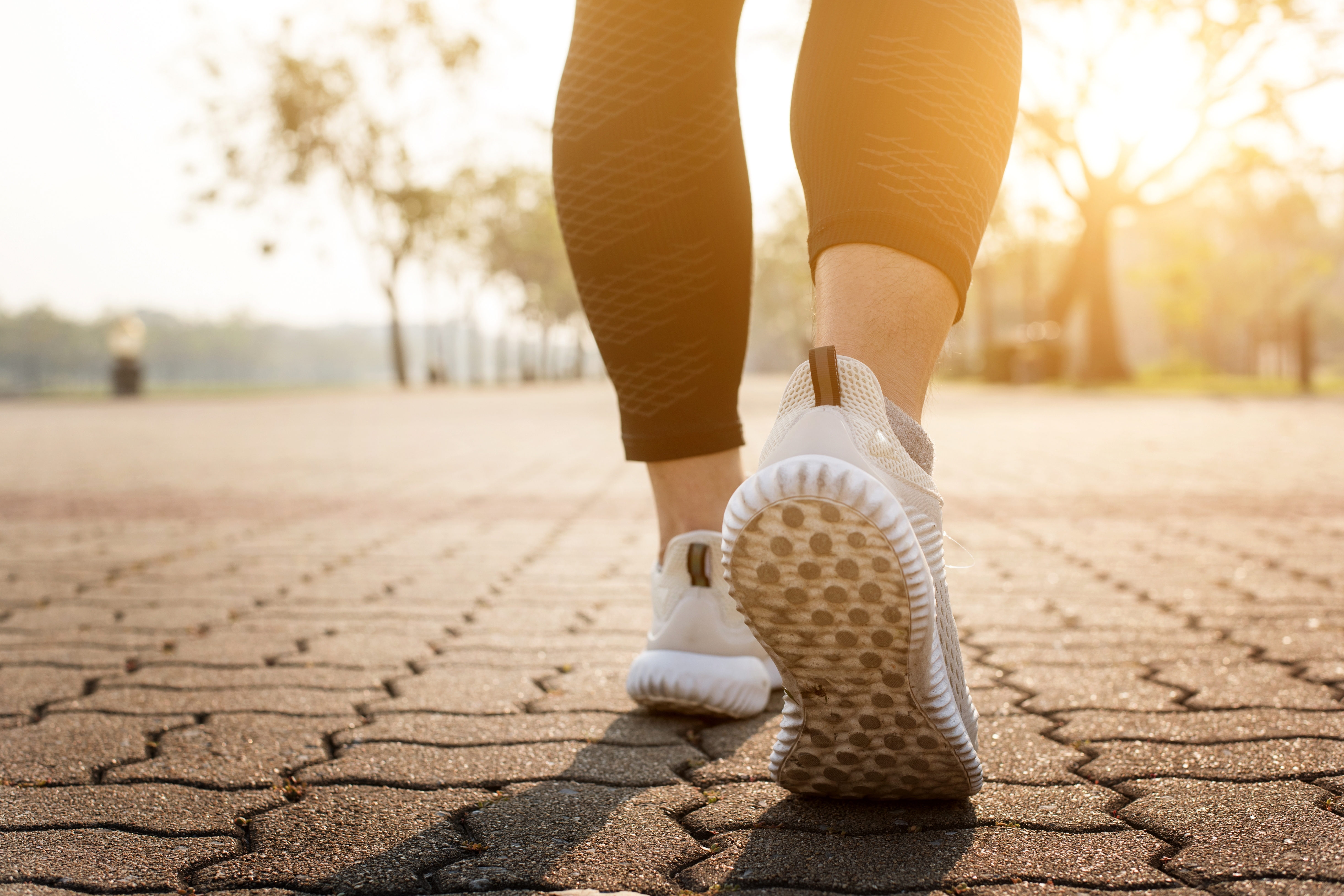 Person walking on a paved path wearing athletic shoes and leggings, captured from behind with a sunny, blurred background.