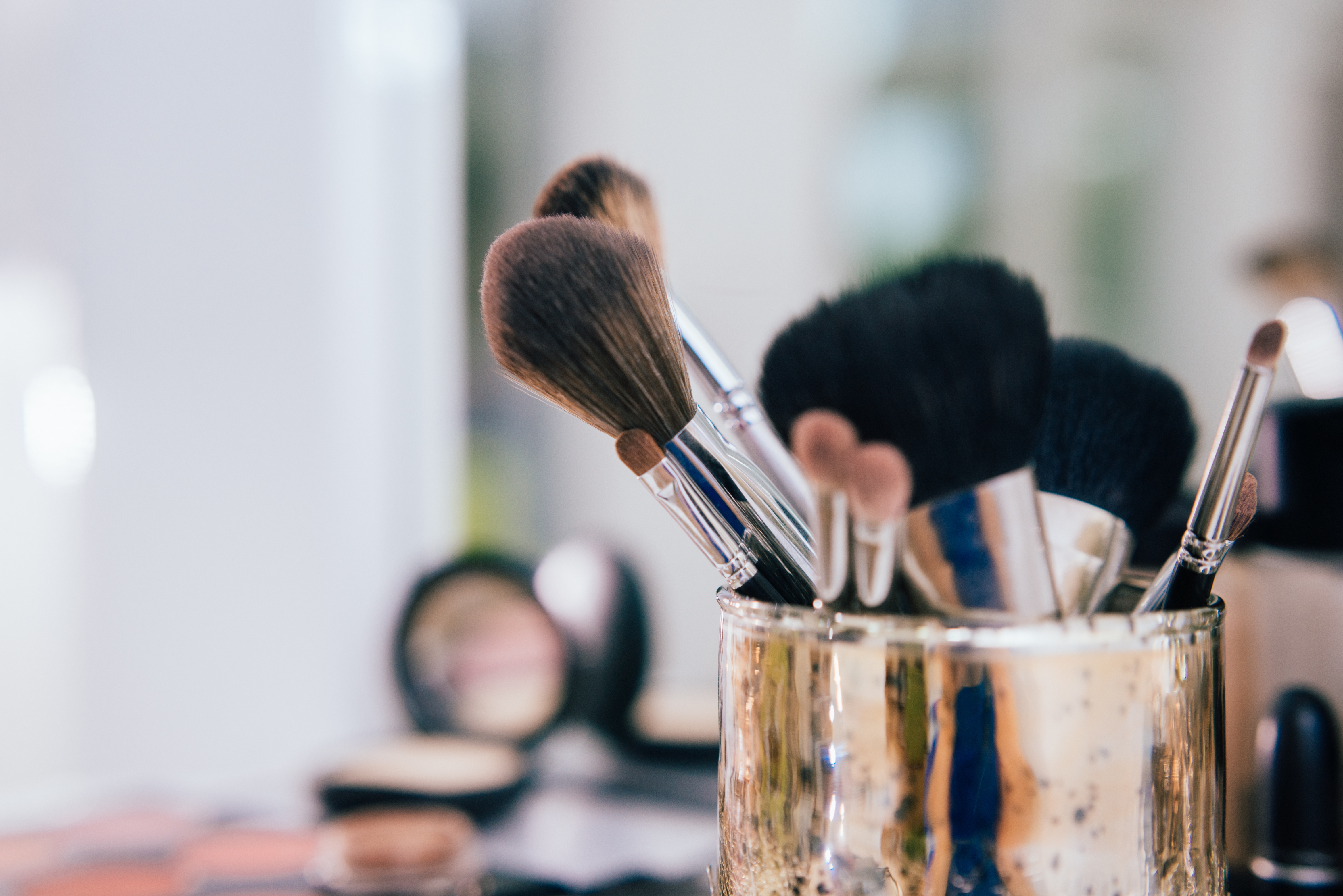 Makeup brushes in a gold holder on a vanity, blurred background with various makeup items visible