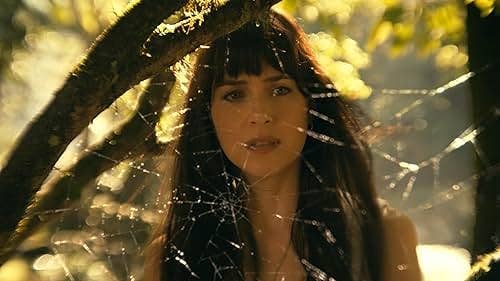 A woman with long hair looks through a spiderweb in a forest setting