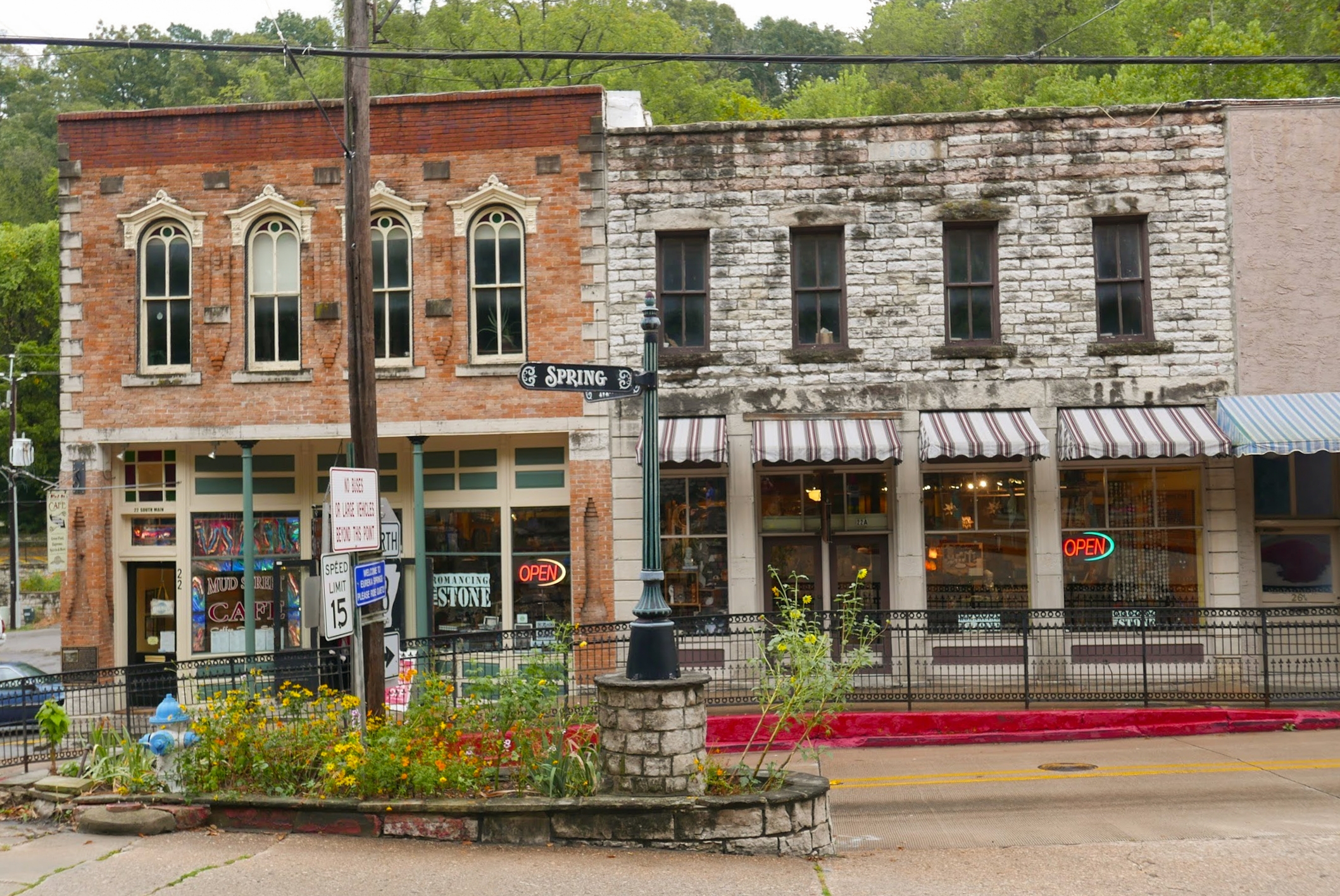 Historic street with two small shops, one in a brick building and one in stone, both with "Open" signs, and a flowerbed in front