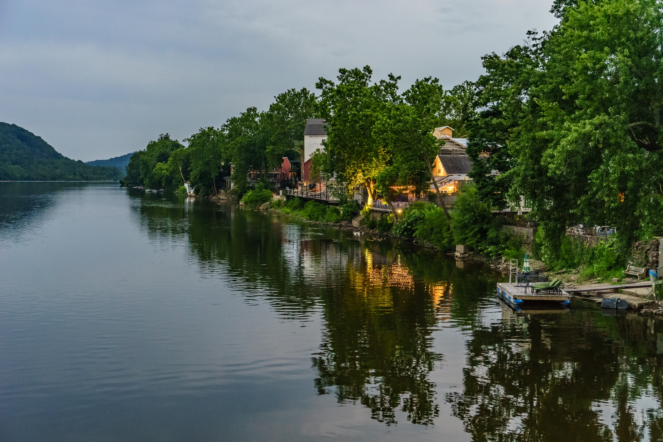 Tree-lined riverbank with small houses and a dock, reflecting on the water. Peaceful and serene landscape scene
